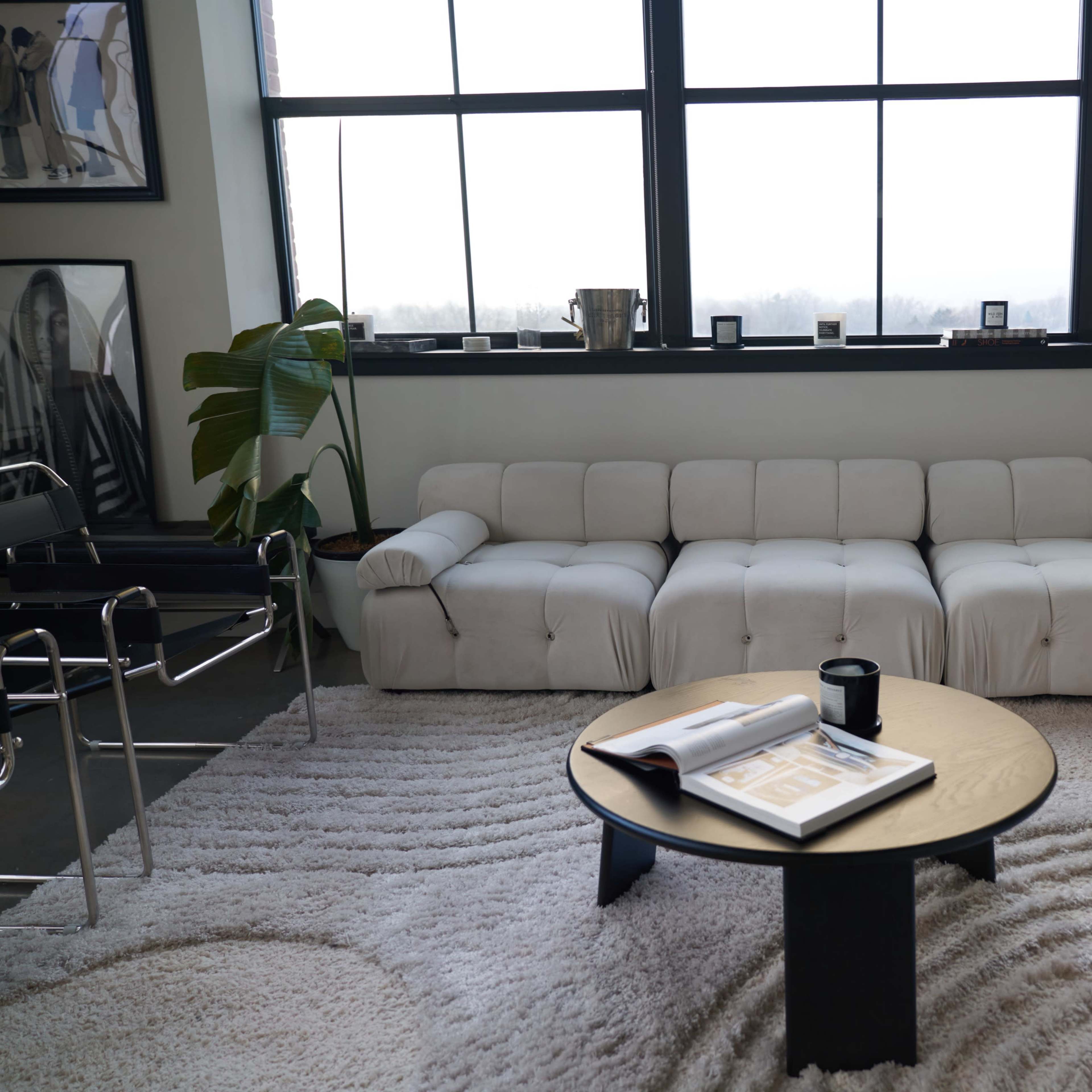 A white sectional sofa is positioned in front of large windows, with a round coffee table and a patterned rug nearby.