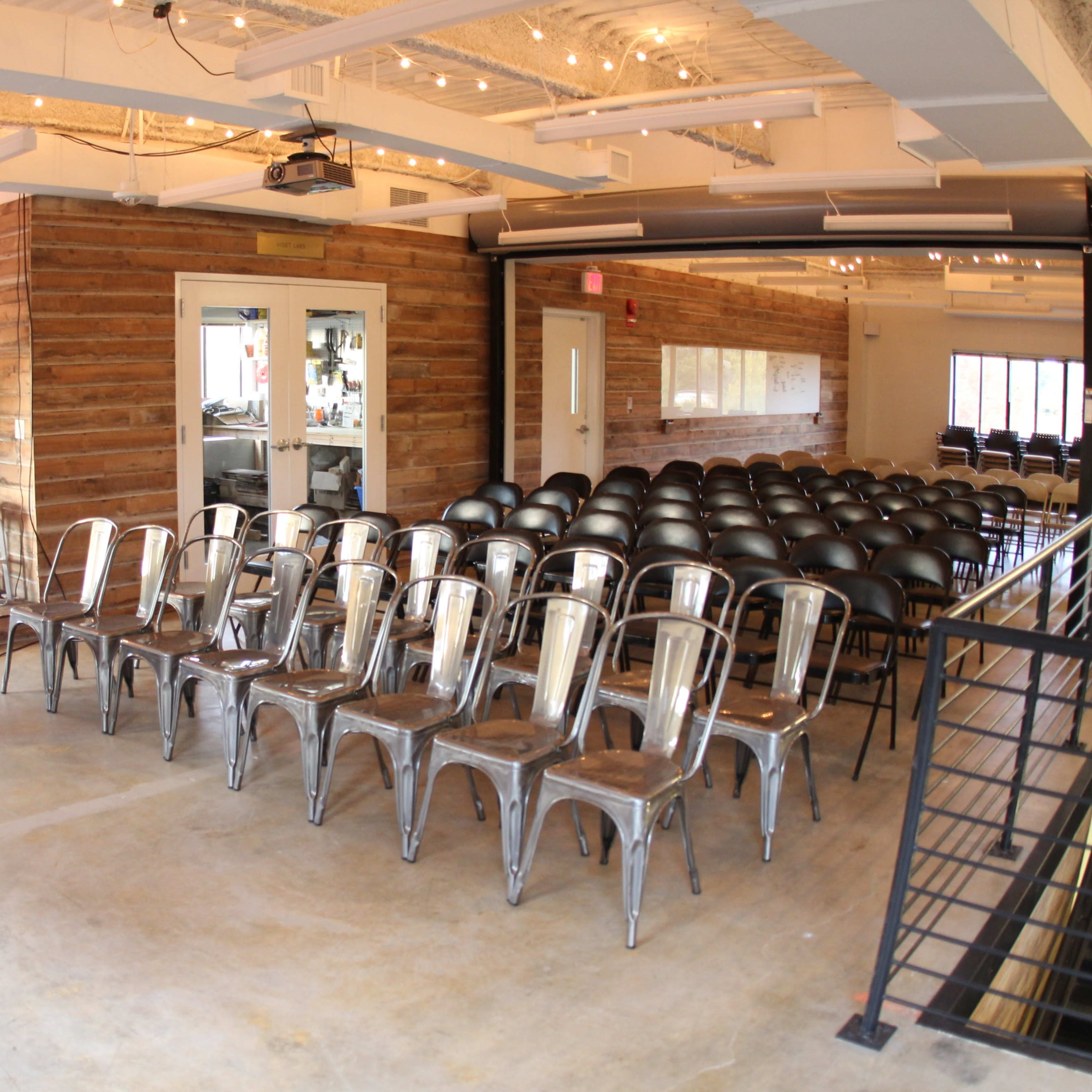A room with a wooden stage and rows of metal chairs set up for an event, featuring large windows and modern decor.