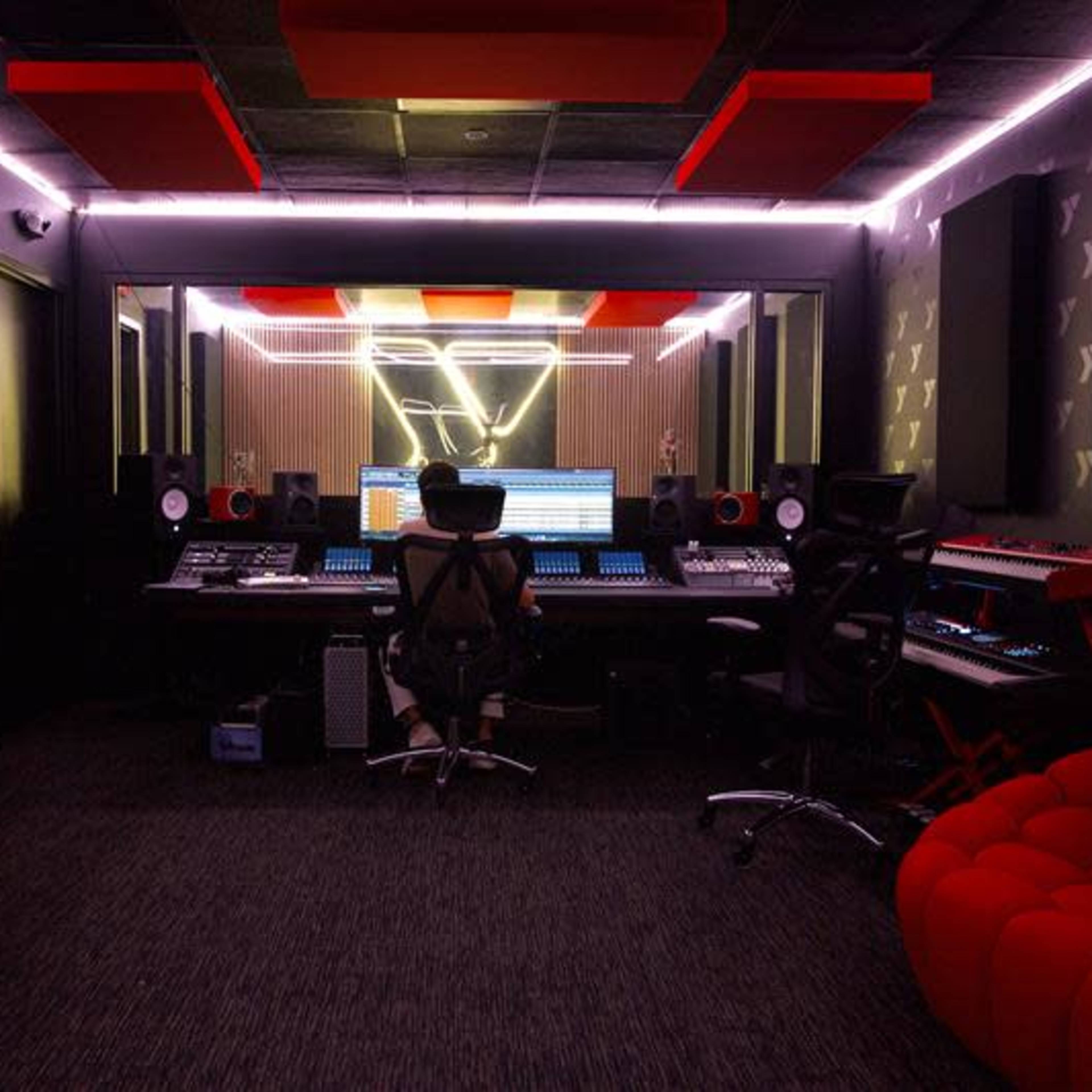 A person sits at a large mixing console in a recording studio, surrounded by various audio equipment and illuminated by neon lighting.
