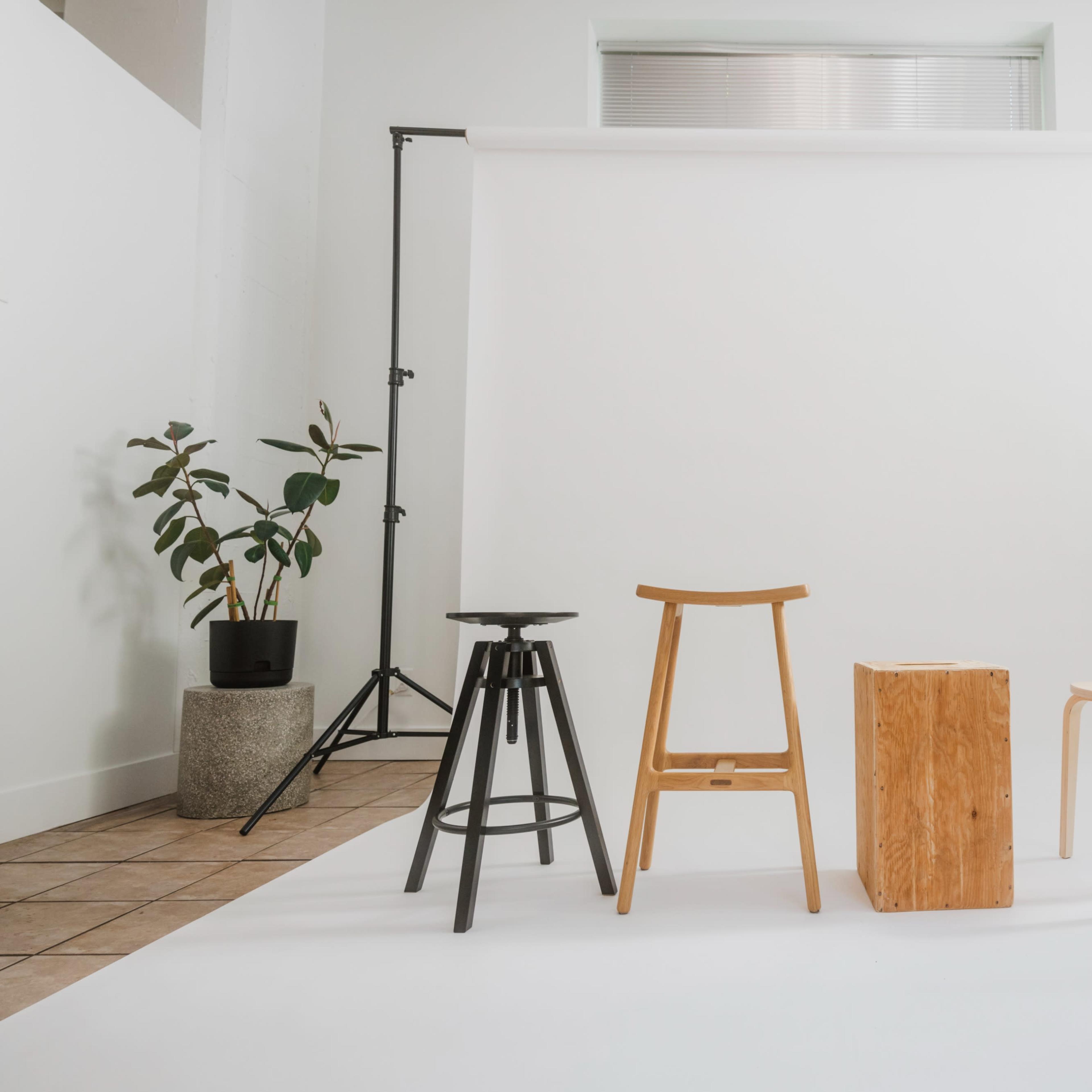 A photo studio setup features three different stools arranged in front of a white backdrop, with potted plants in the corners.