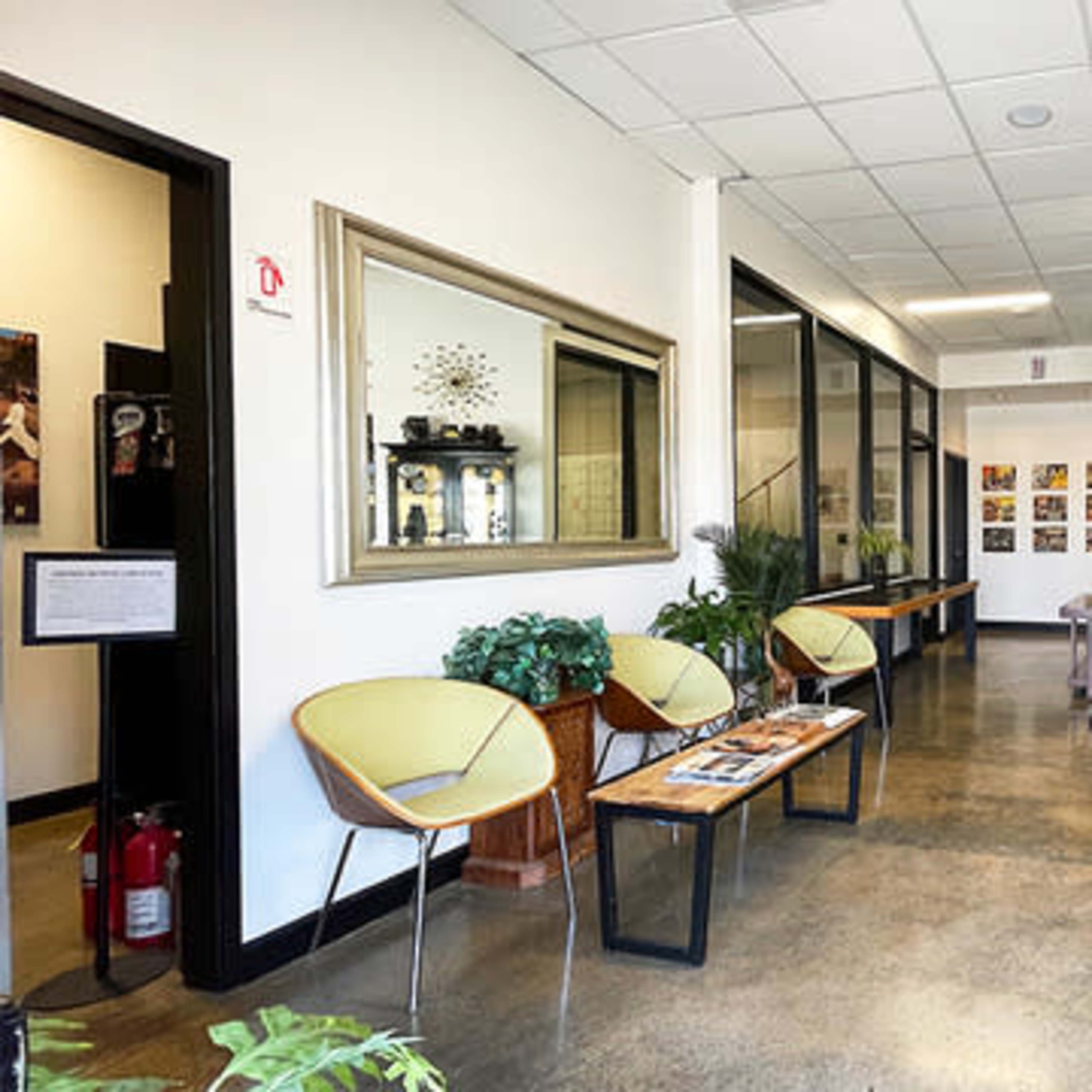 A modern waiting area with two seating areas, decorative plants, framed artwork on the walls, and a black cabinet displaying various items.