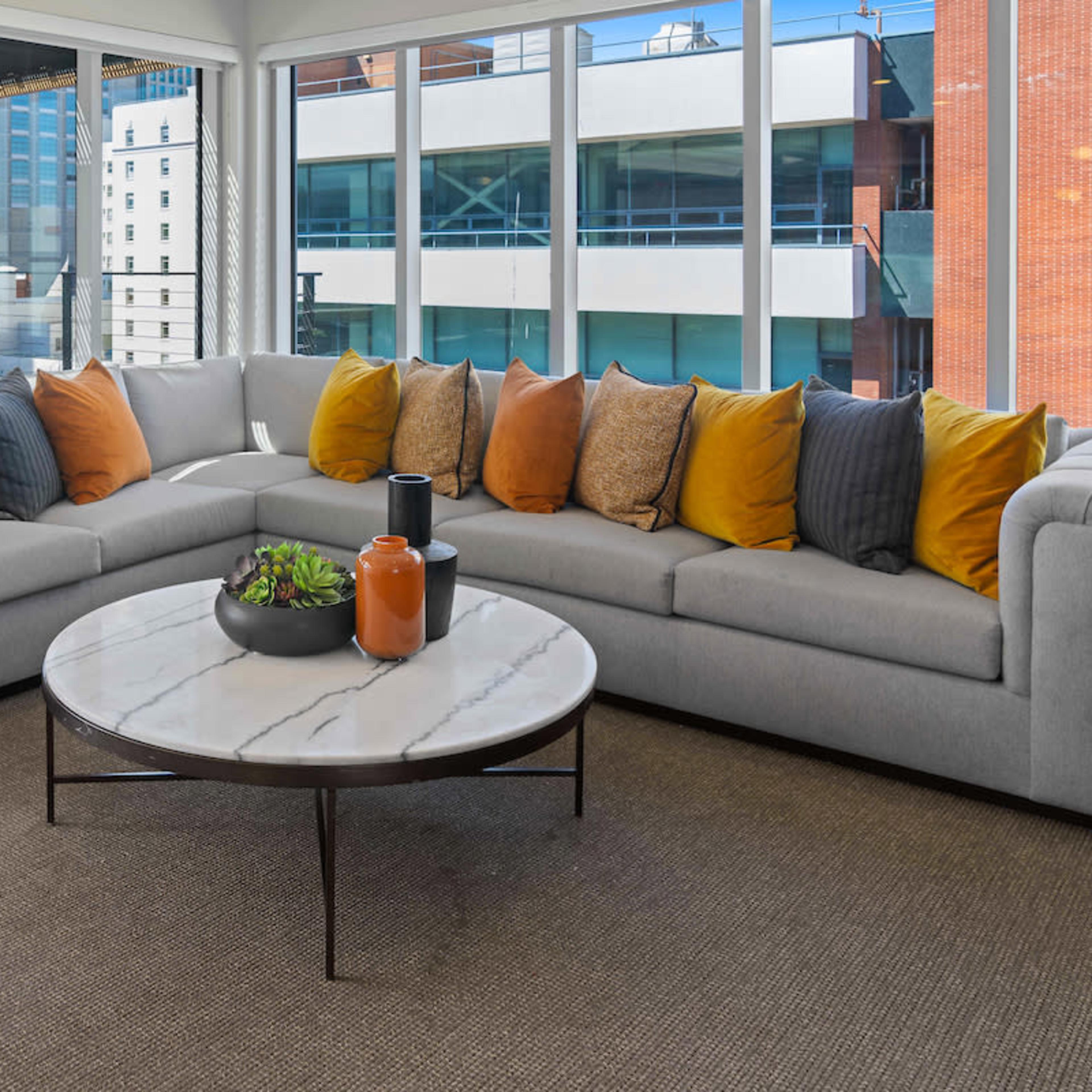 A modern living room features a gray sectional sofa with colorful throw pillows surrounding a round marble coffee table, all set against large windows overlooking an urban landscape.
