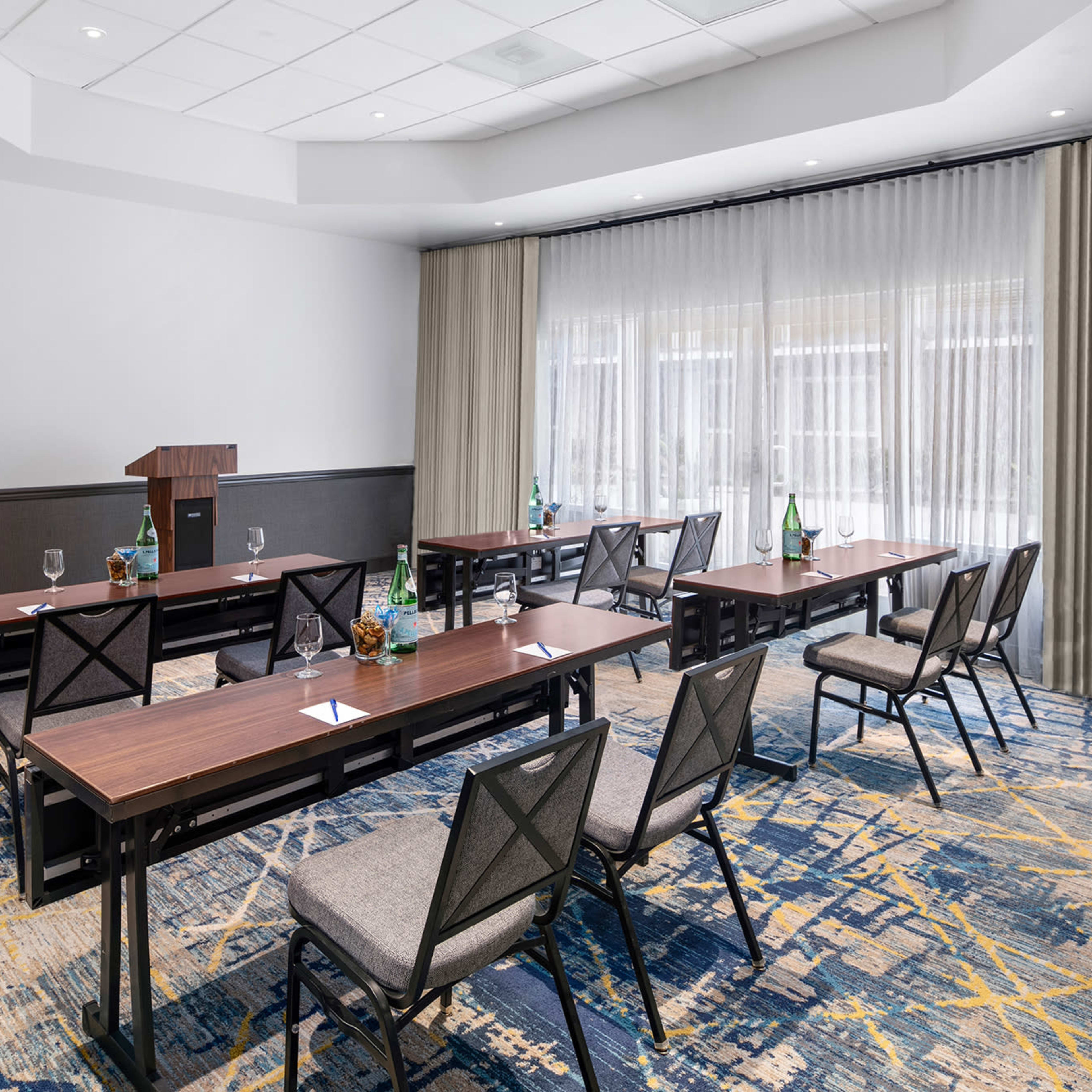 A conference room features several rows of tables with chairs arranged for a meeting, along with a podium and natural light coming through large windows.