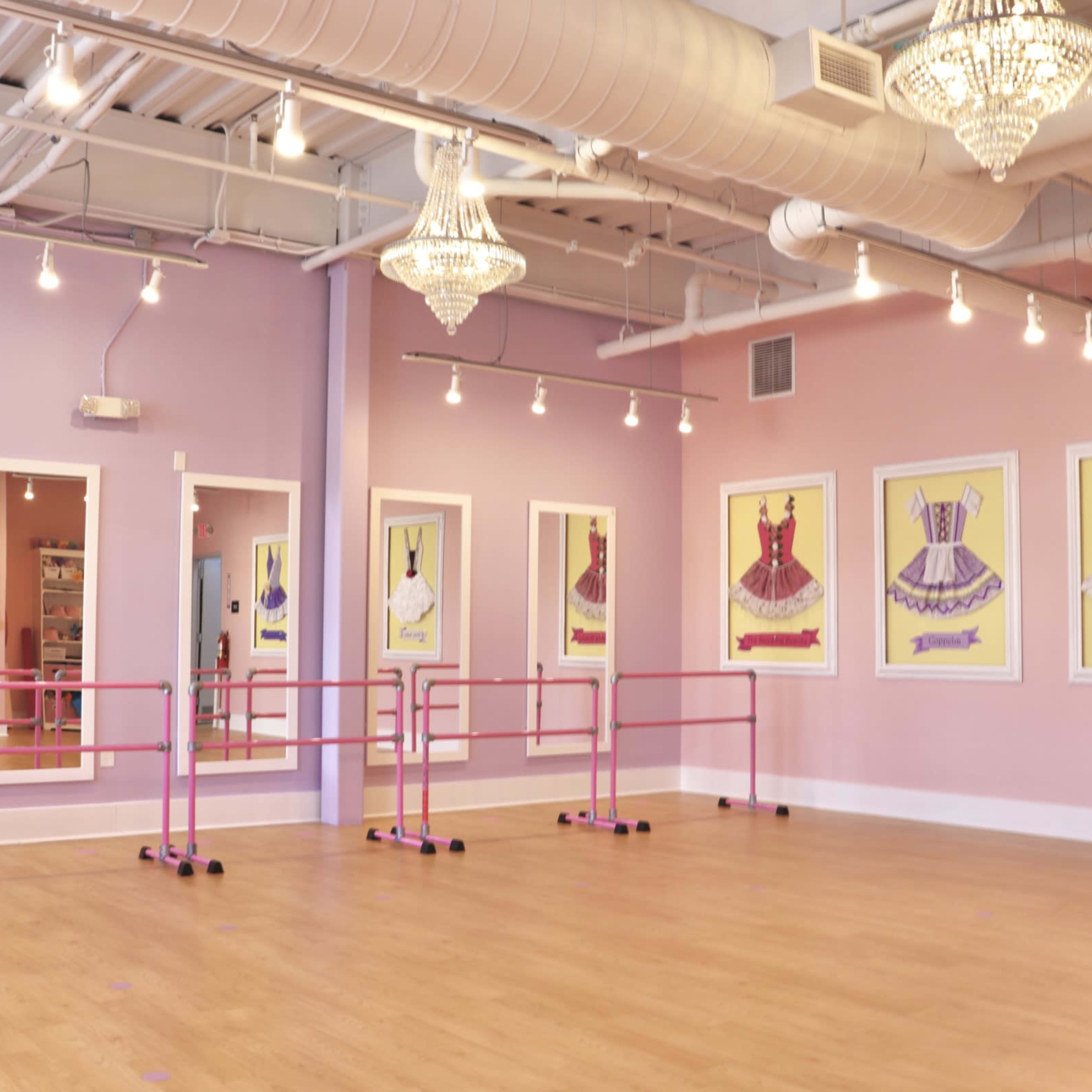 A dance studio with mirrors, pink ballet barres, and framed illustrations of ballet dresses on pastel walls.