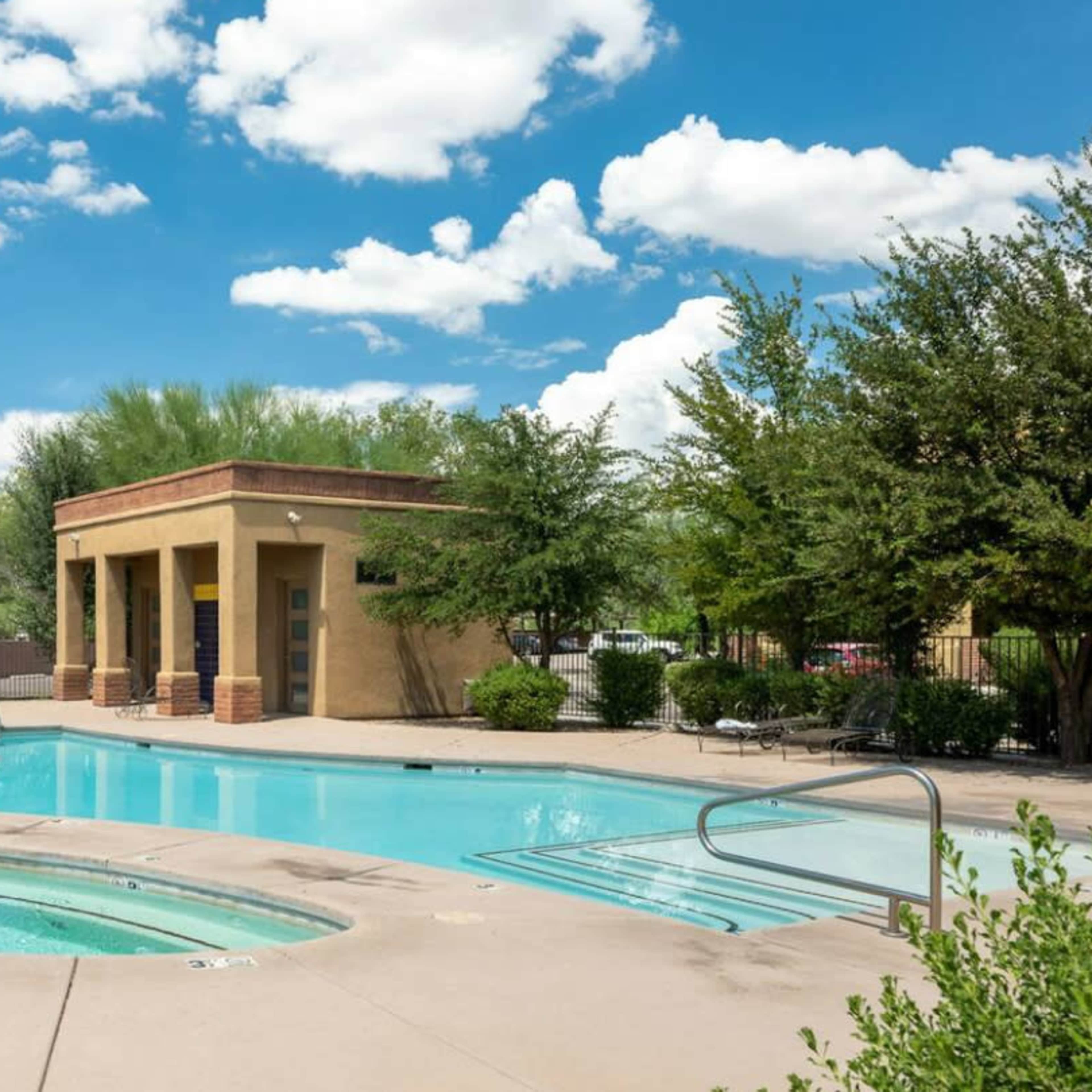 A swimming pool area with a hot tub, surrounded by greenery and a small building in the background under a blue sky with clouds.