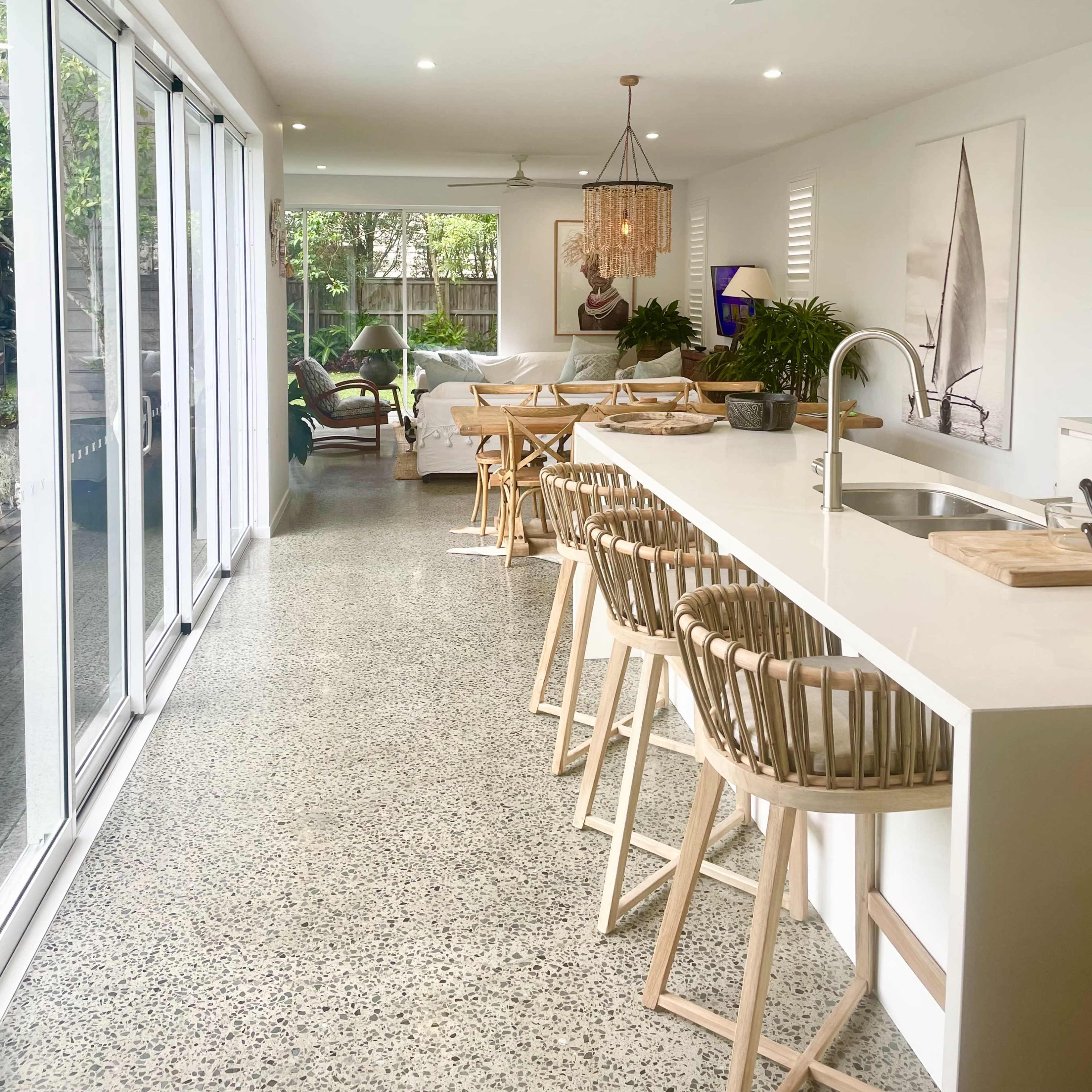 A modern kitchen with a white island bar and wooden stools opens to a living area with large windows and contemporary furnishings.