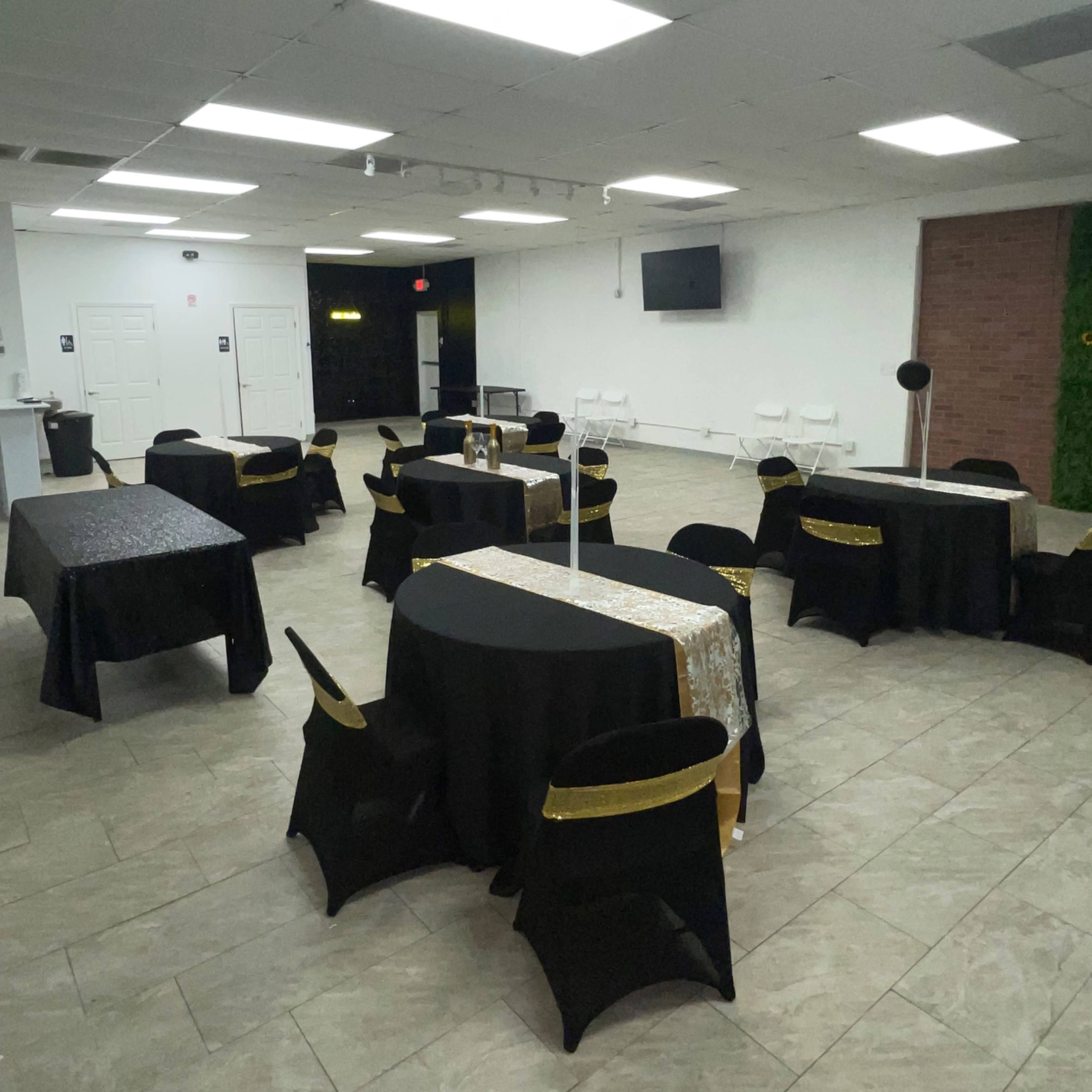 A room set up for an event, featuring tables covered in black and gold tablecloths arranged throughout the space with white chairs nearby.