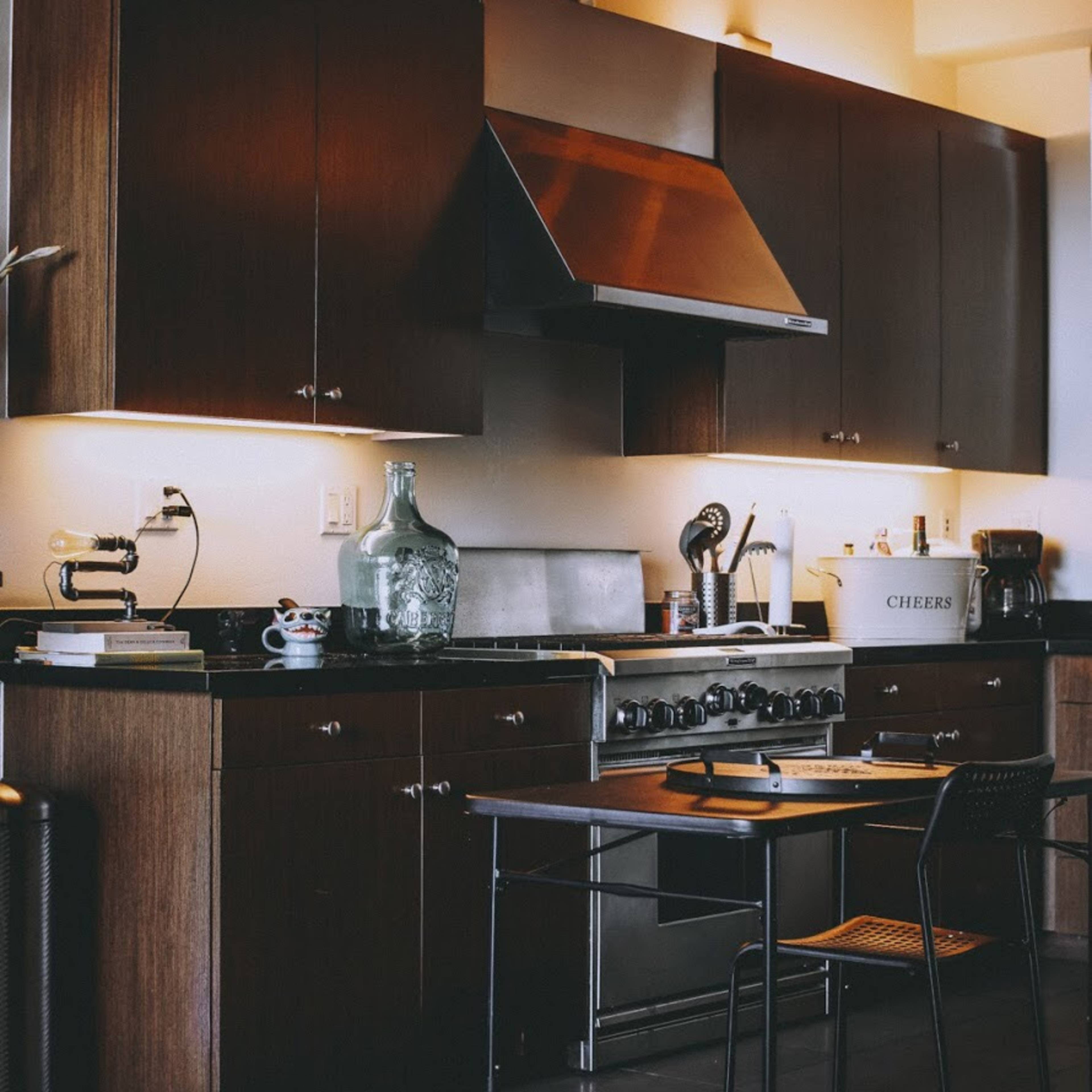 A modern kitchen features dark wooden cabinets, a stainless steel range hood, and a central stainless steel stove with a glass jug on the counter.