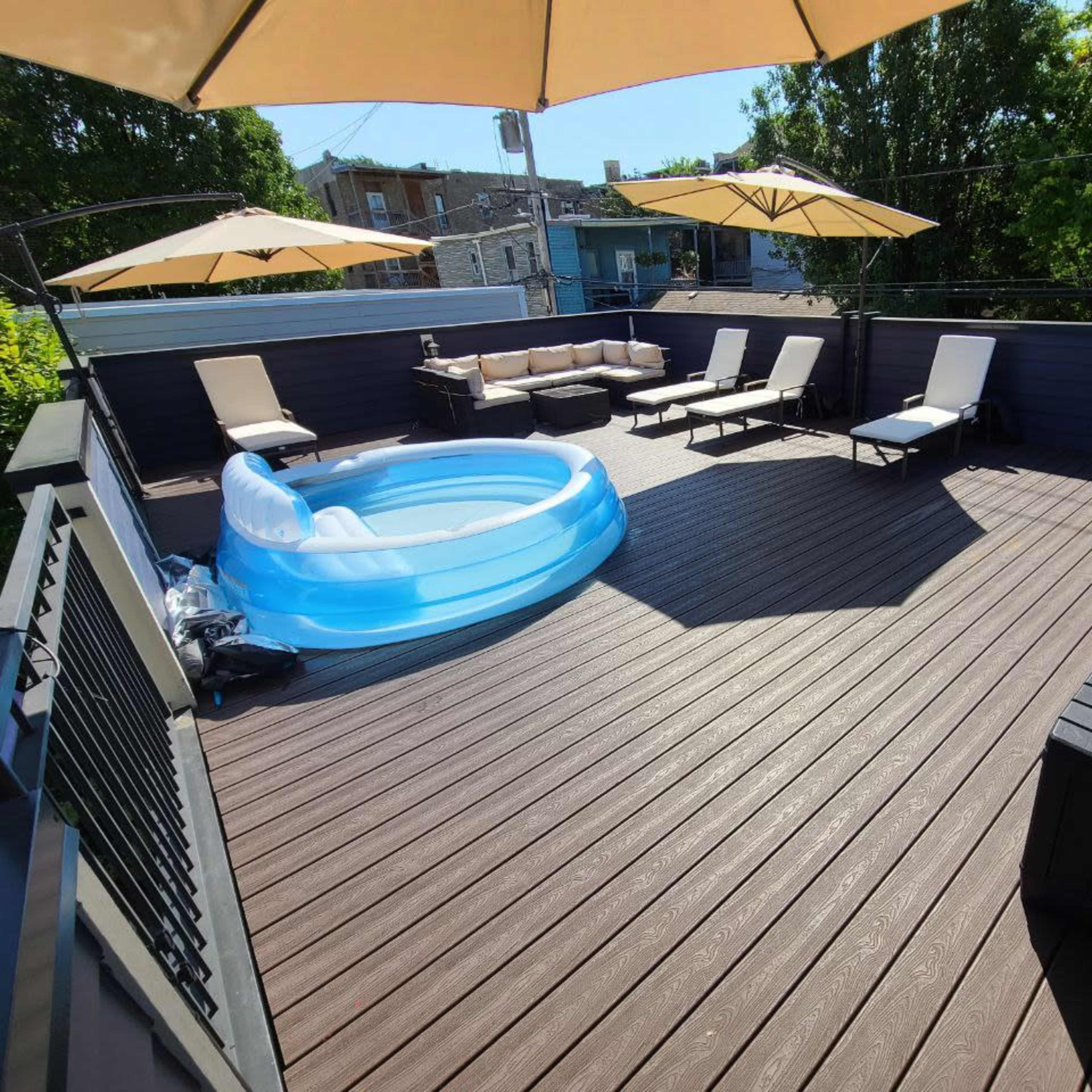 A rooftop terrace featuring a circular inflatable pool, several lounge chairs, and two umbrellas, all set on a wooden deck.