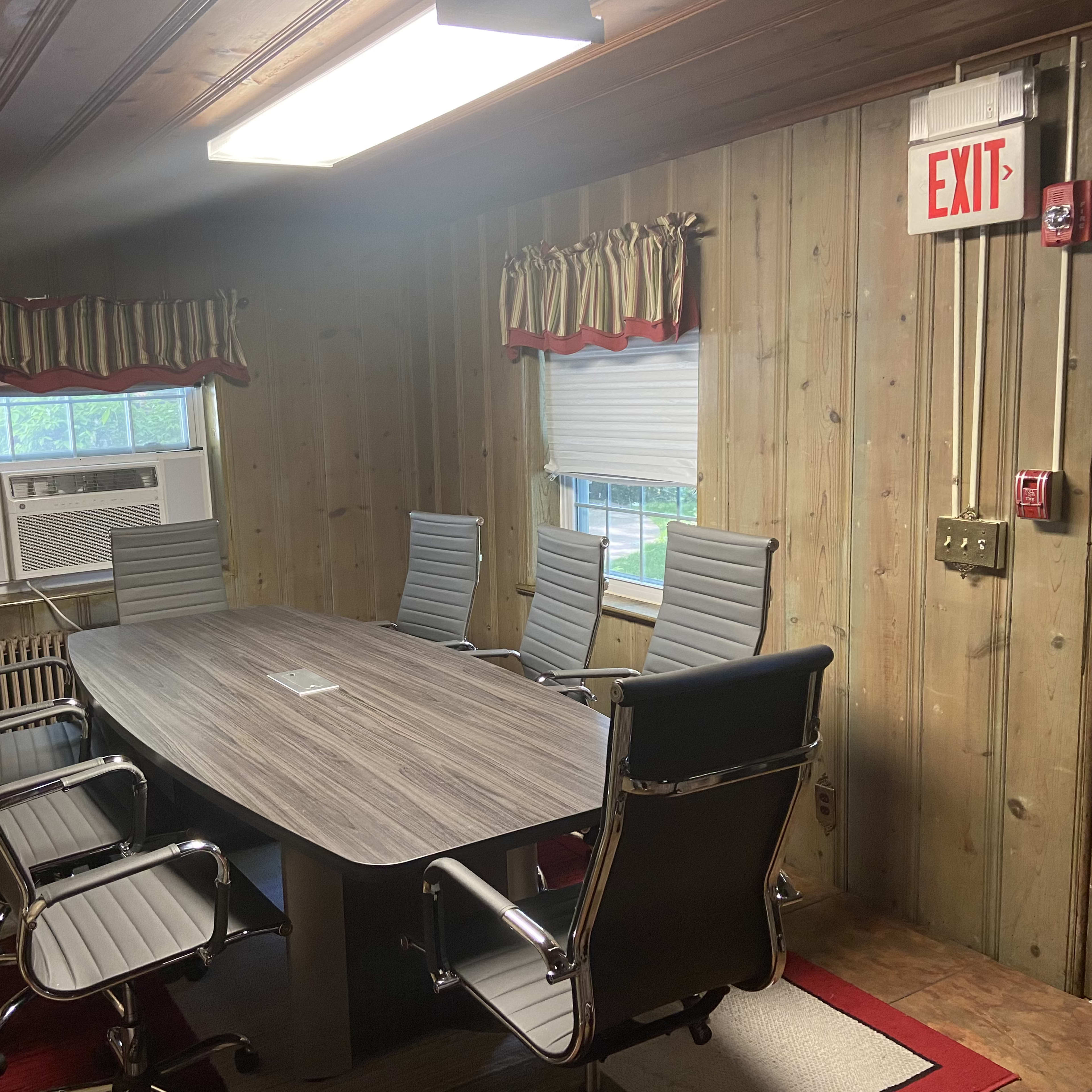 A conference room features a long oval table surrounded by six ergonomic chairs, with wooden paneling on the walls and an air conditioning unit visible in the corner.