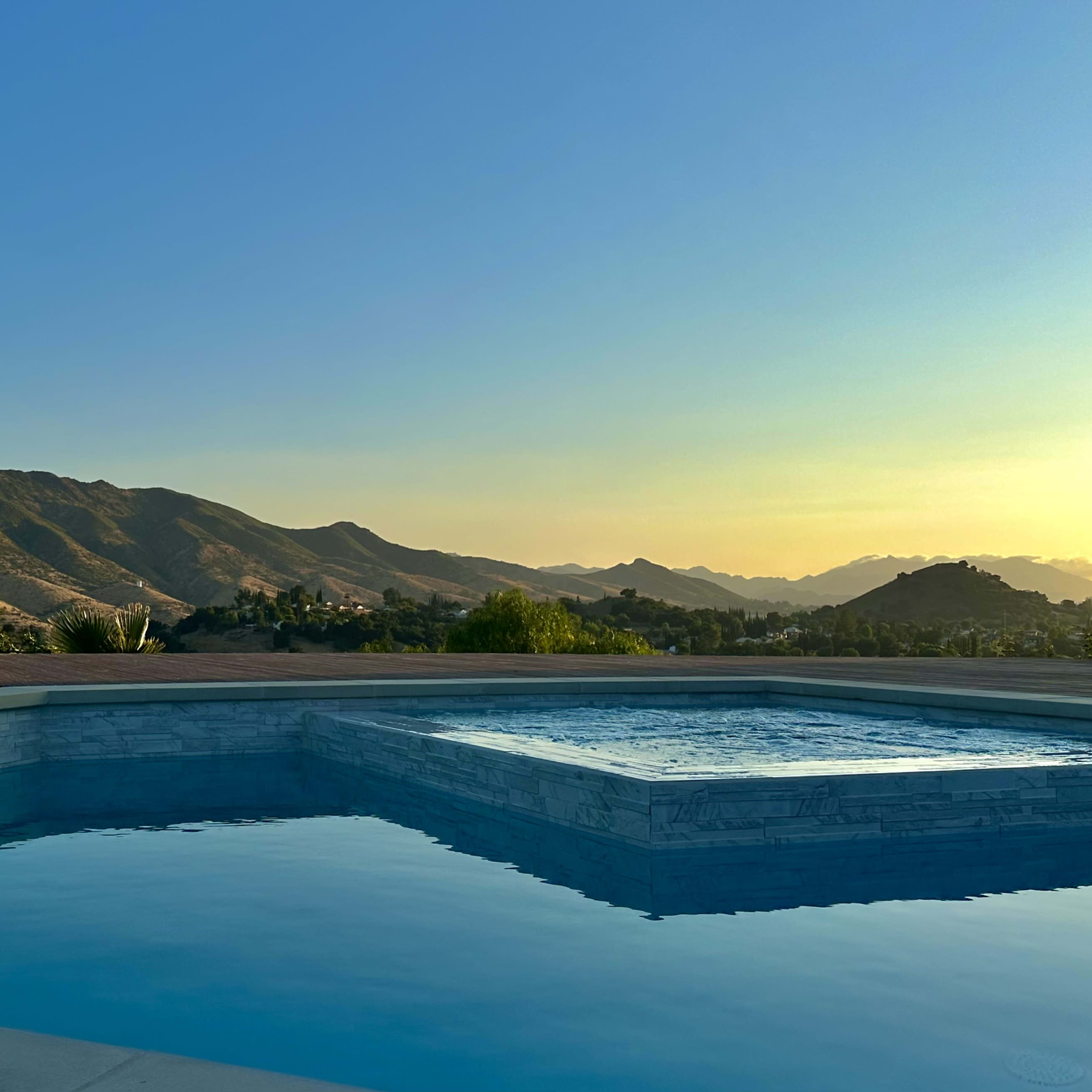 A swimming pool with a spa overlooks a mountainous landscape under a clear sky at sunset.