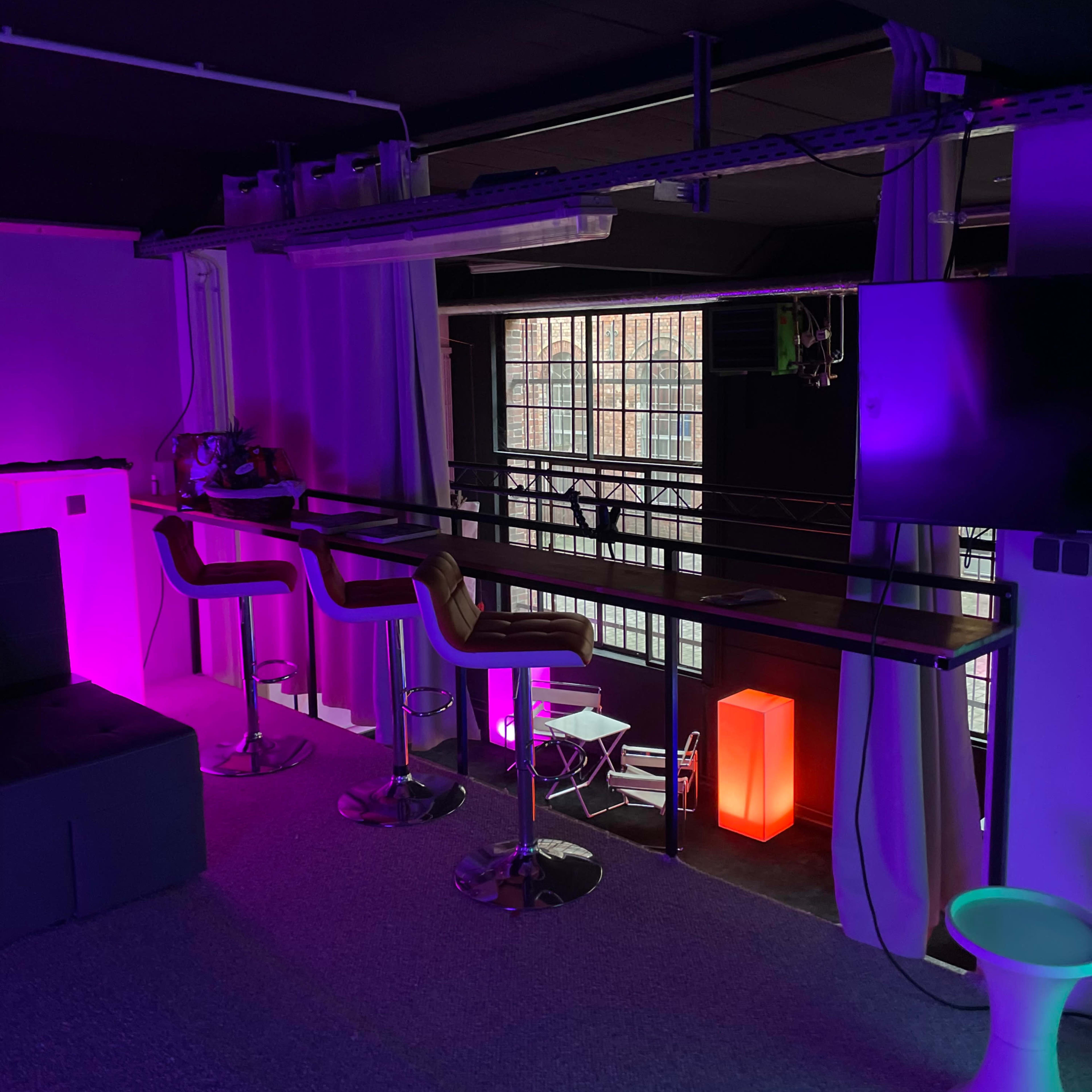 A dimly lit room featuring modern barstools by a counter, illuminated by purple and red lights, with large windows in the background.