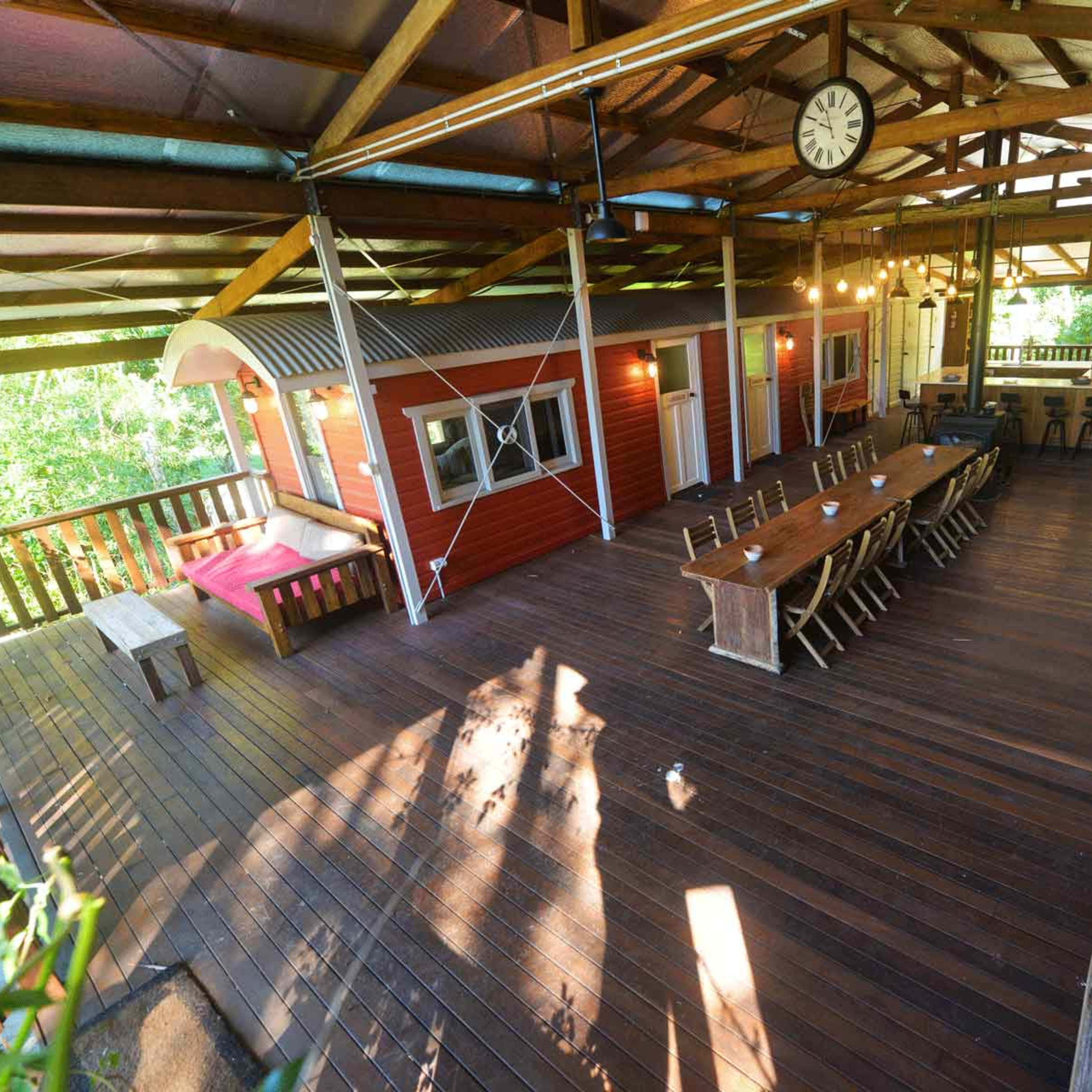 A spacious wooden deck with several seating areas, including a long dining table and a smaller seating arrangement, all beneath a covered structure supported by wooden beams.