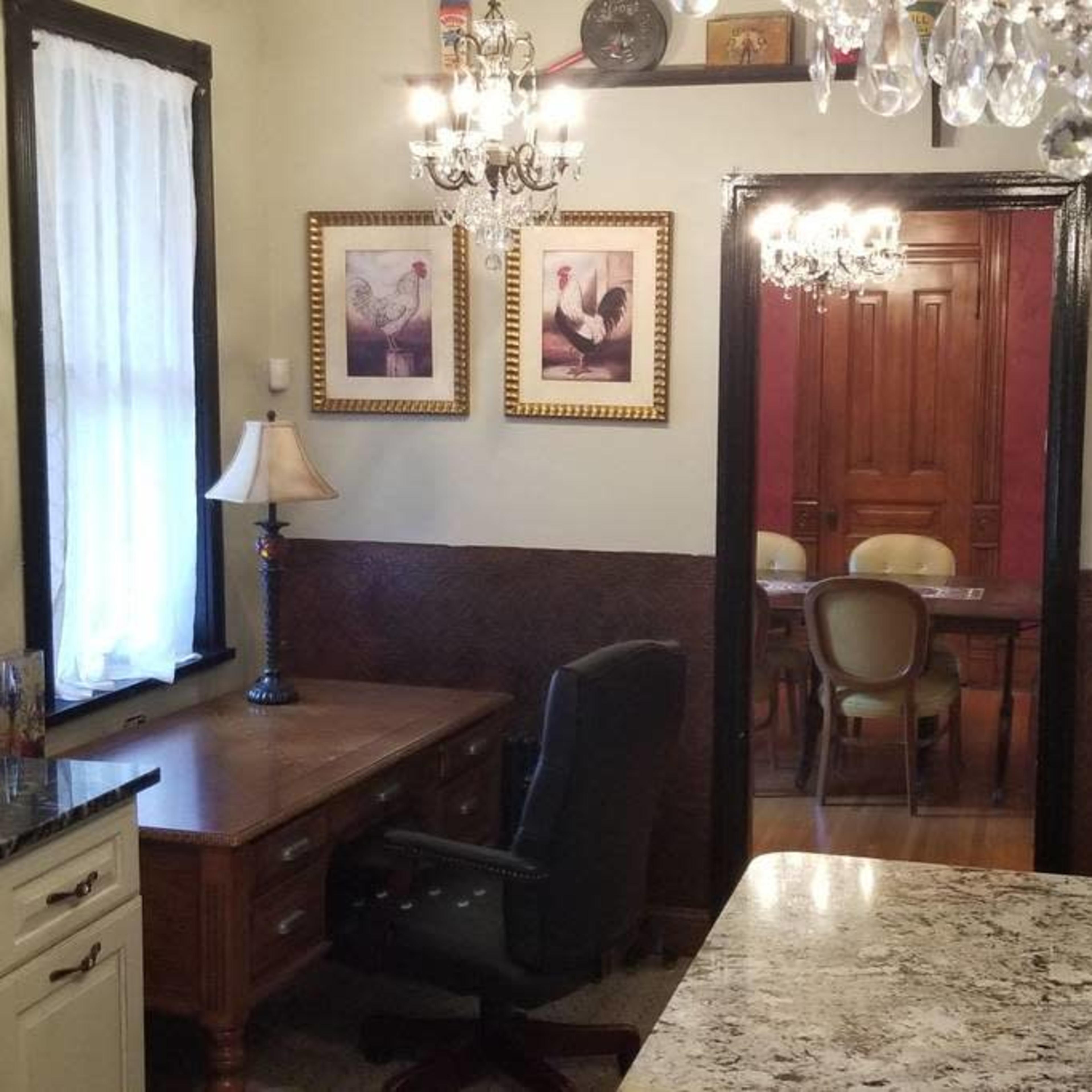 A kitchen with a desk and chair in the foreground, leading to a dining area visible through an open doorway, with framed pictures on the walls and chandeliers overhead.