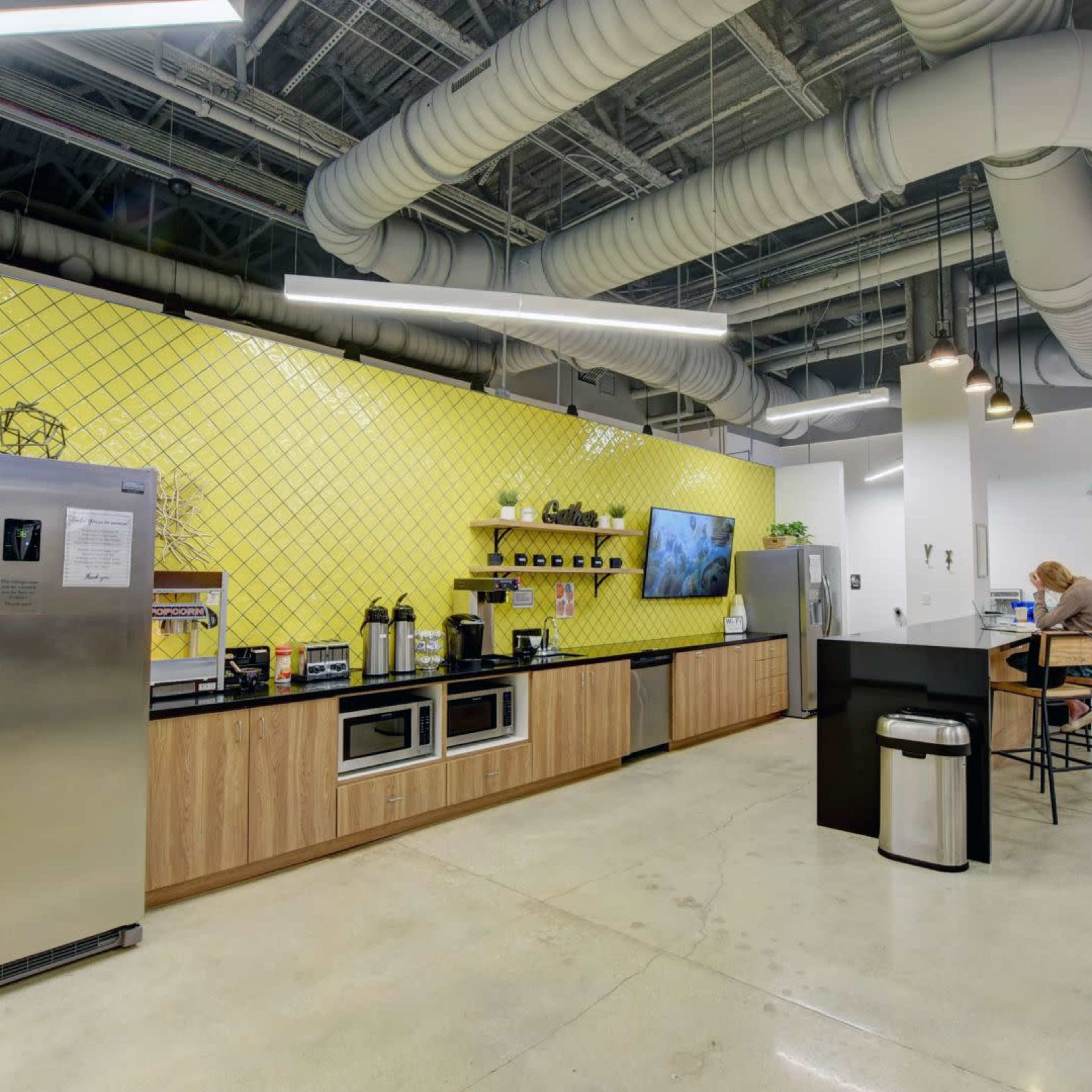 A modern kitchen area in an office, featuring a stainless steel refrigerator, coffee machines, and a large yellow tiled wall with a television above a wooden counter.