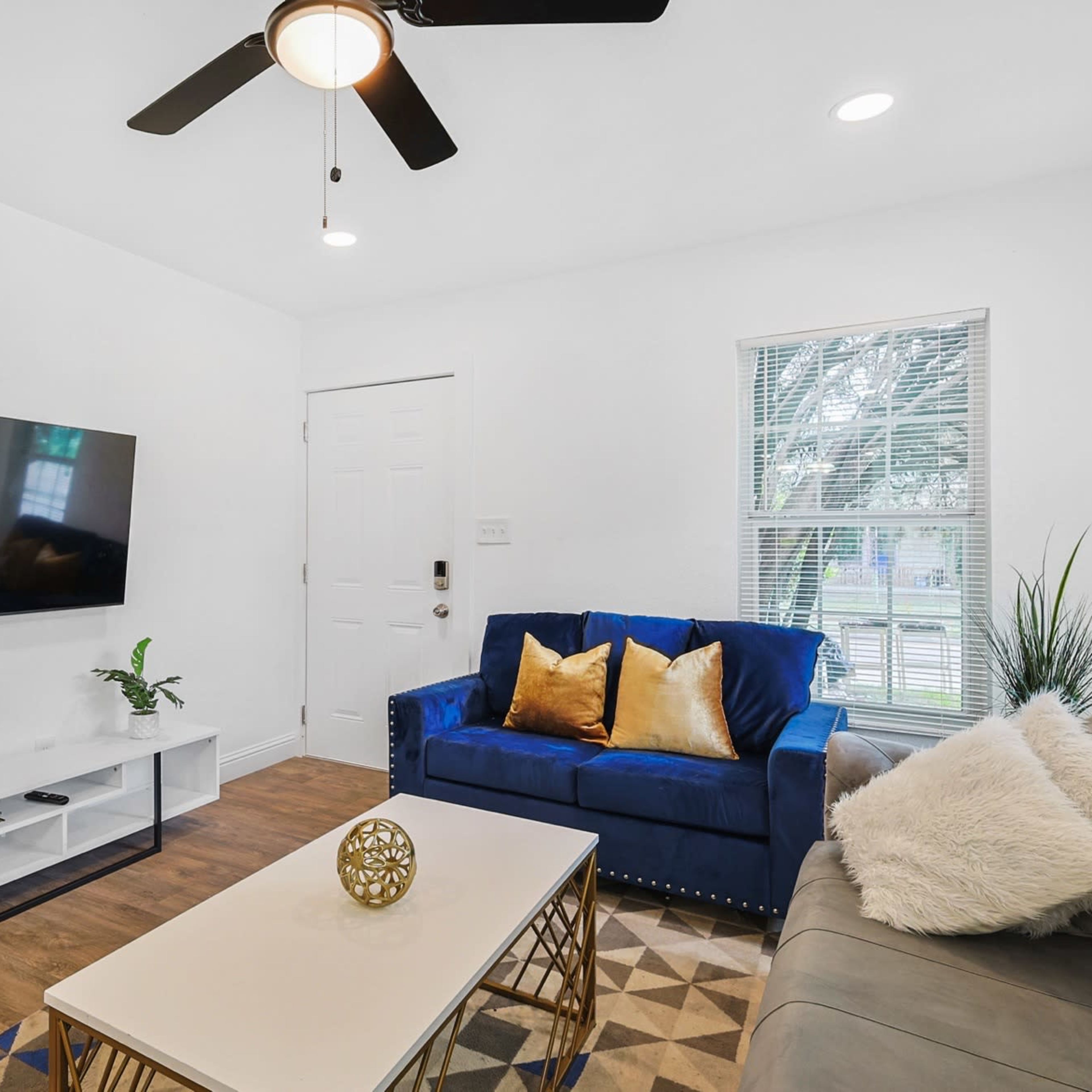 A living room features a blue sofa, a light-colored coffee table, a mounted television, and plants.