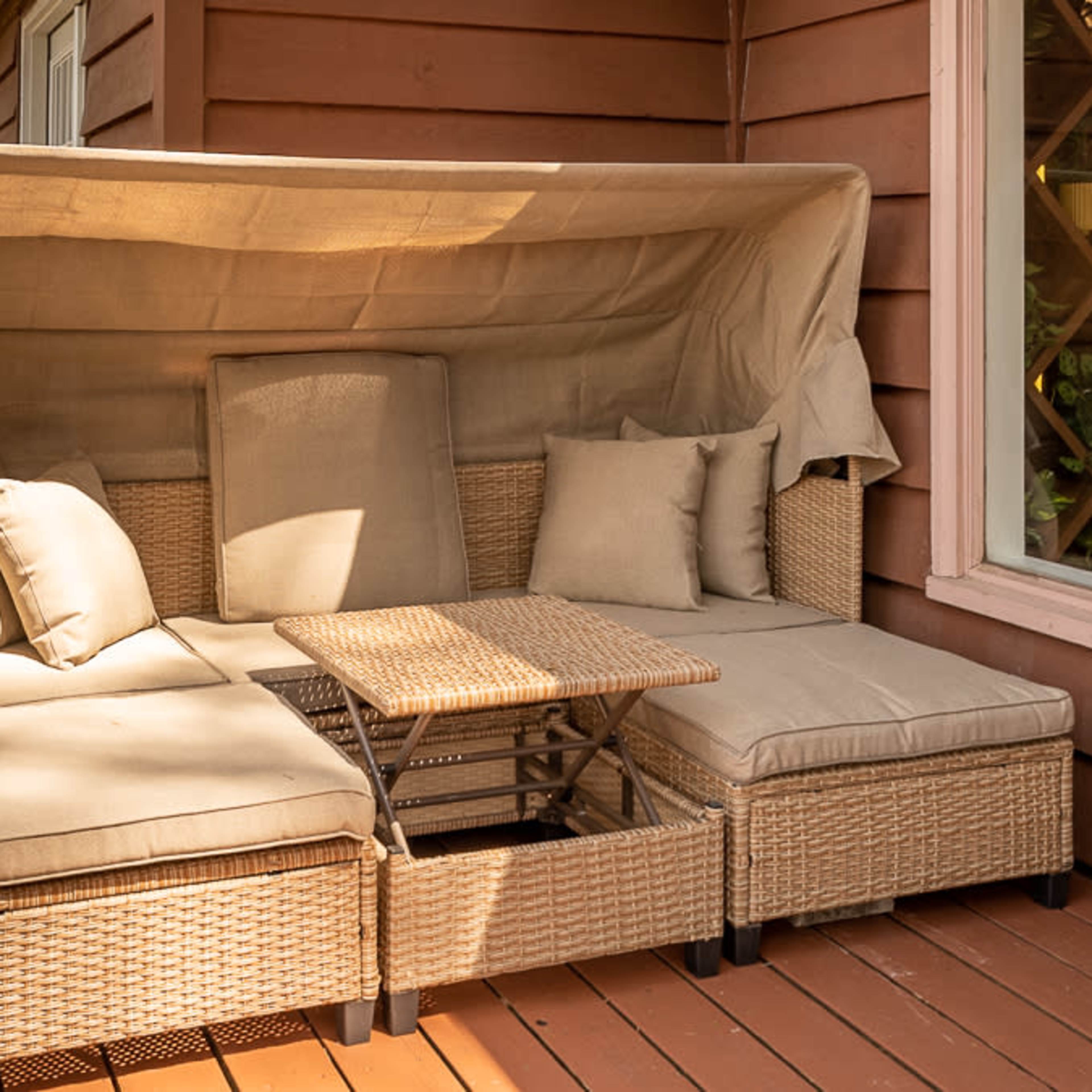 A covered outdoor seating area with a sectional sofa and a small table is situated on a wooden deck next to a house.