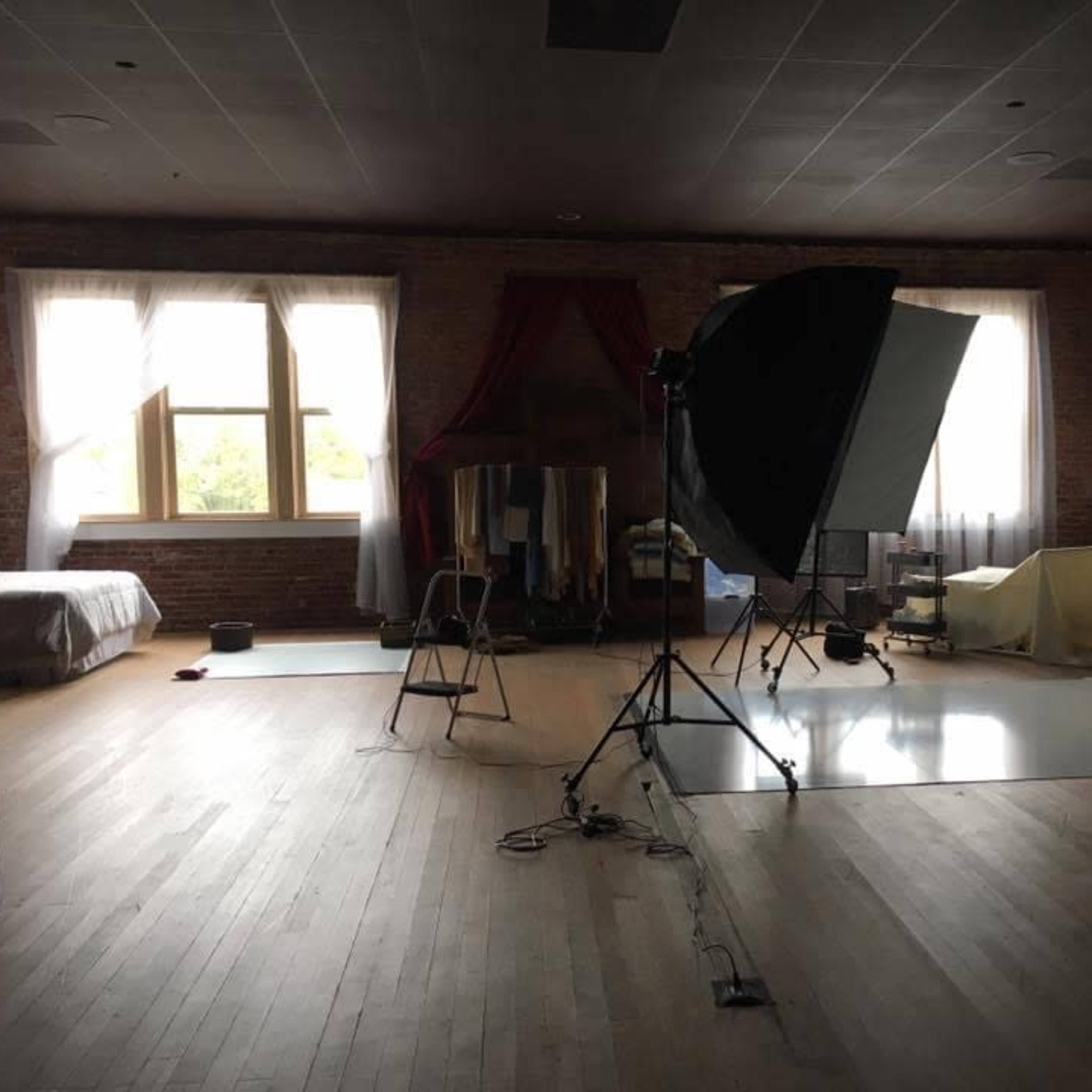 A photography studio with a large softbox, reflective materials, and a bed, set against exposed brick walls and large windows.