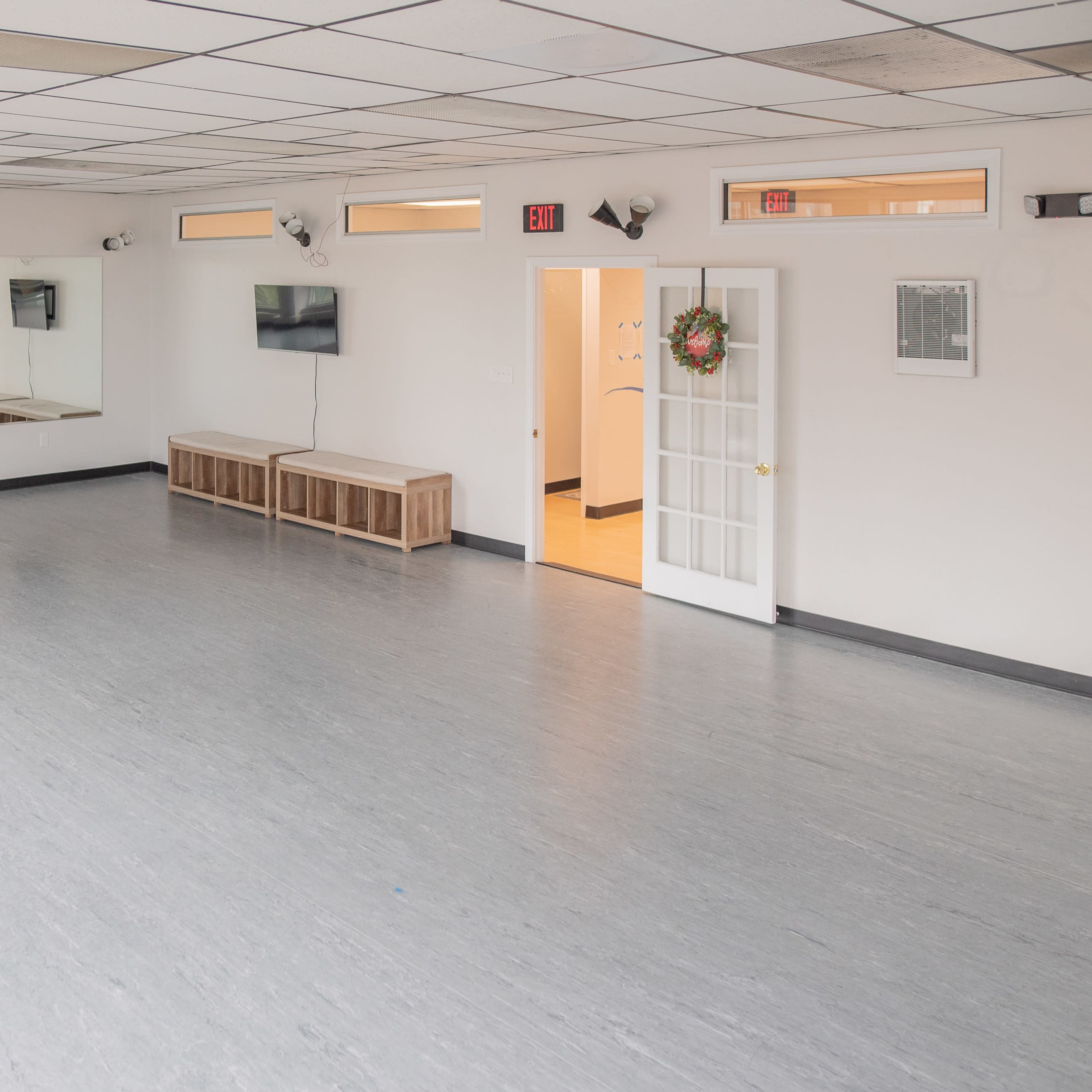 A spacious, empty studio with wooden benches, a TV on the wall, and a door leading to a hallway.