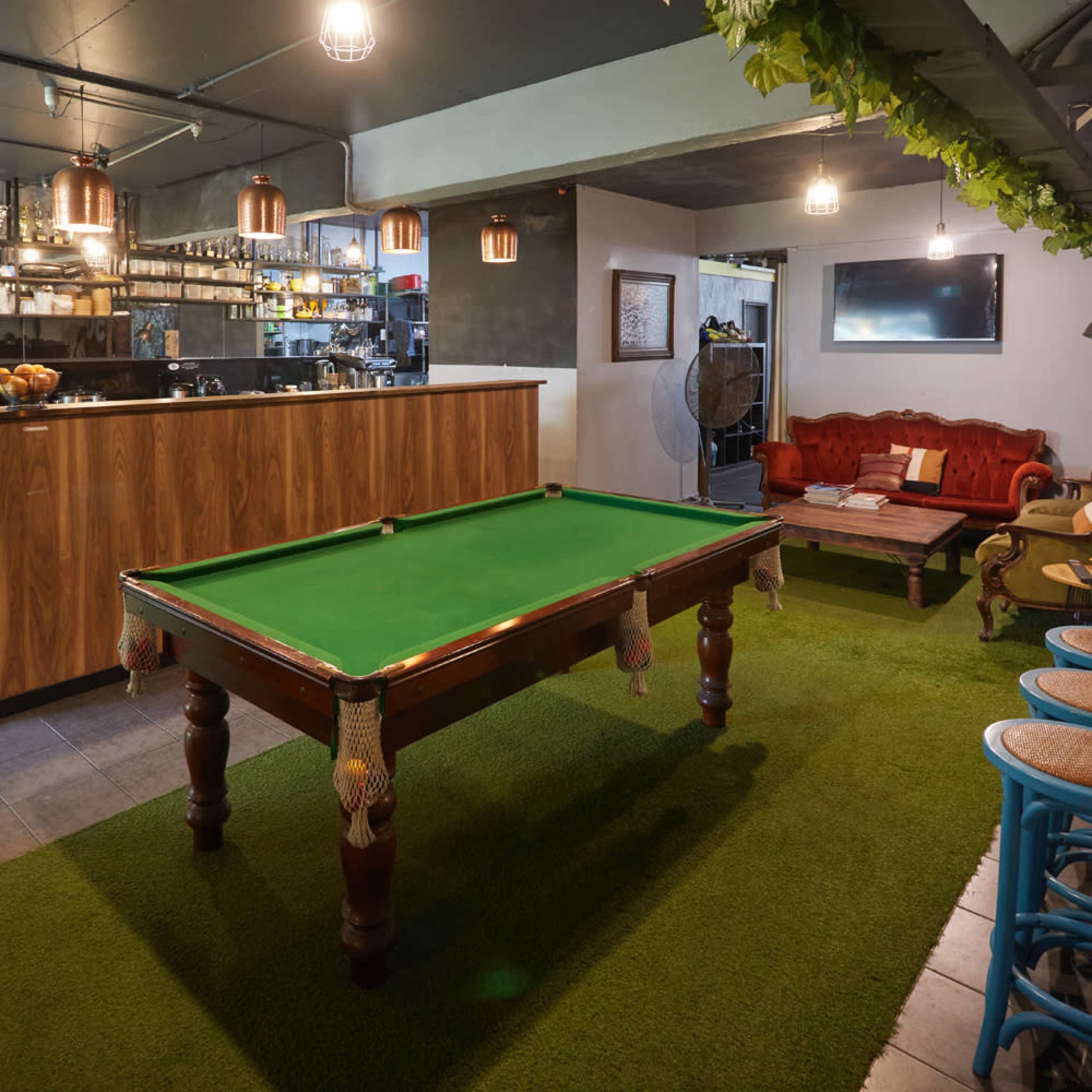 A cozy lounge area featuring a green billiard table, a bar counter with shelves, and a seating arrangement with red and wooden chairs on a grass-like carpet.