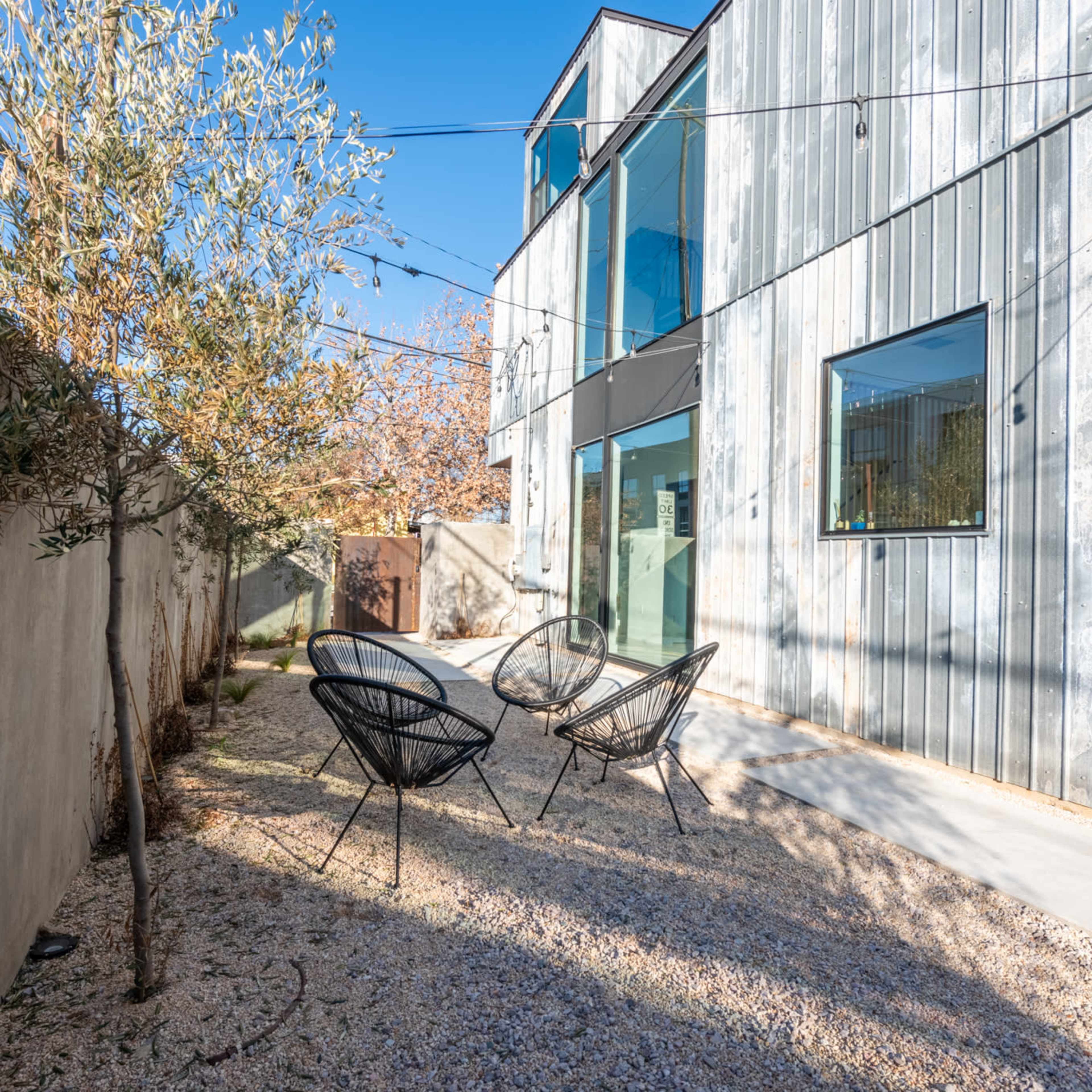 A landscaped outdoor space features three black chairs on gravel, surrounded by walls and a modern building with large windows.