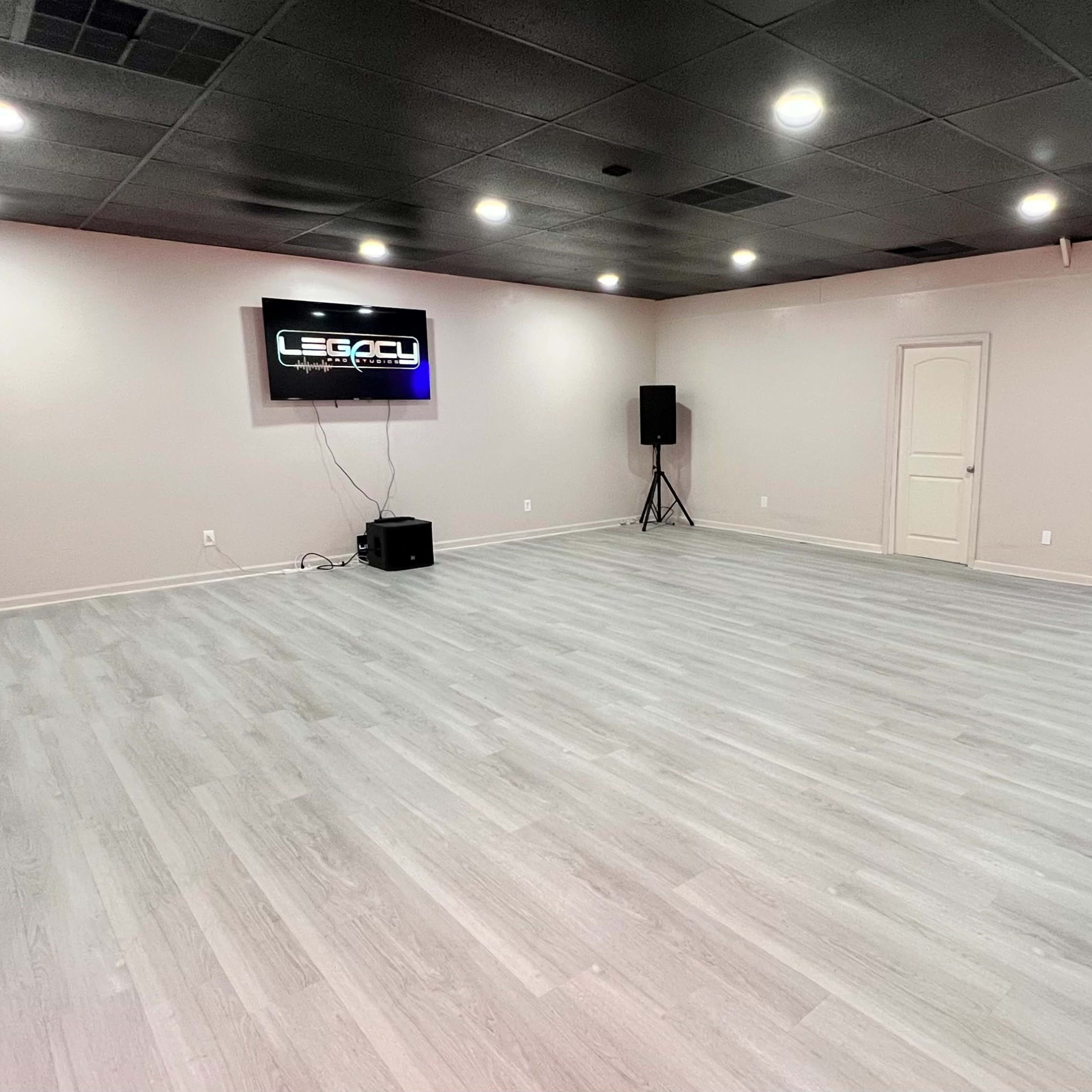 An empty room with gray wooden flooring, a wall-mounted screen displaying the word "LEGACY," two floor speakers, and a set of double doors leading outside.
