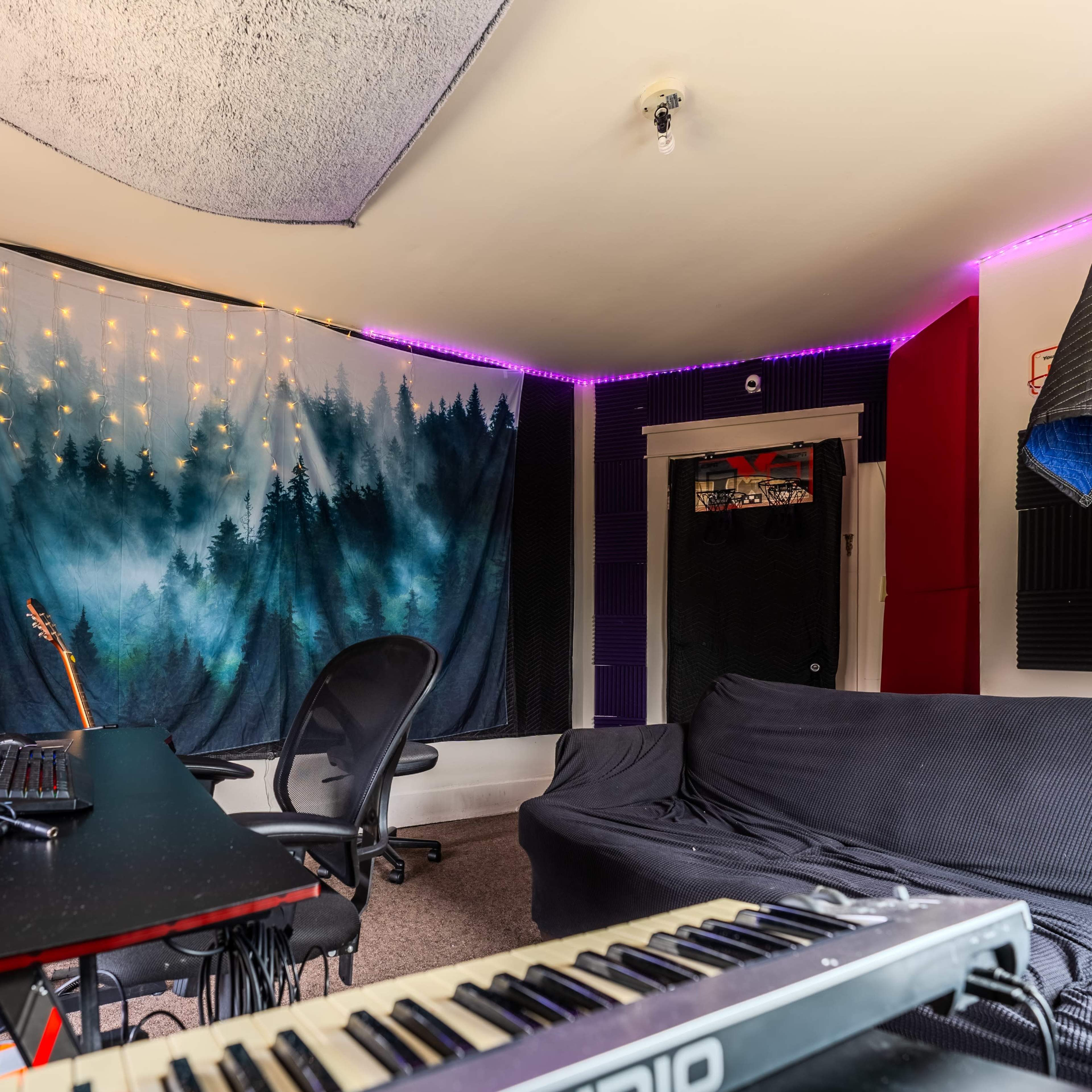 A home studio features a keyboard, a desk with two monitors, and a wall adorned with a forest backdrop and string lights.