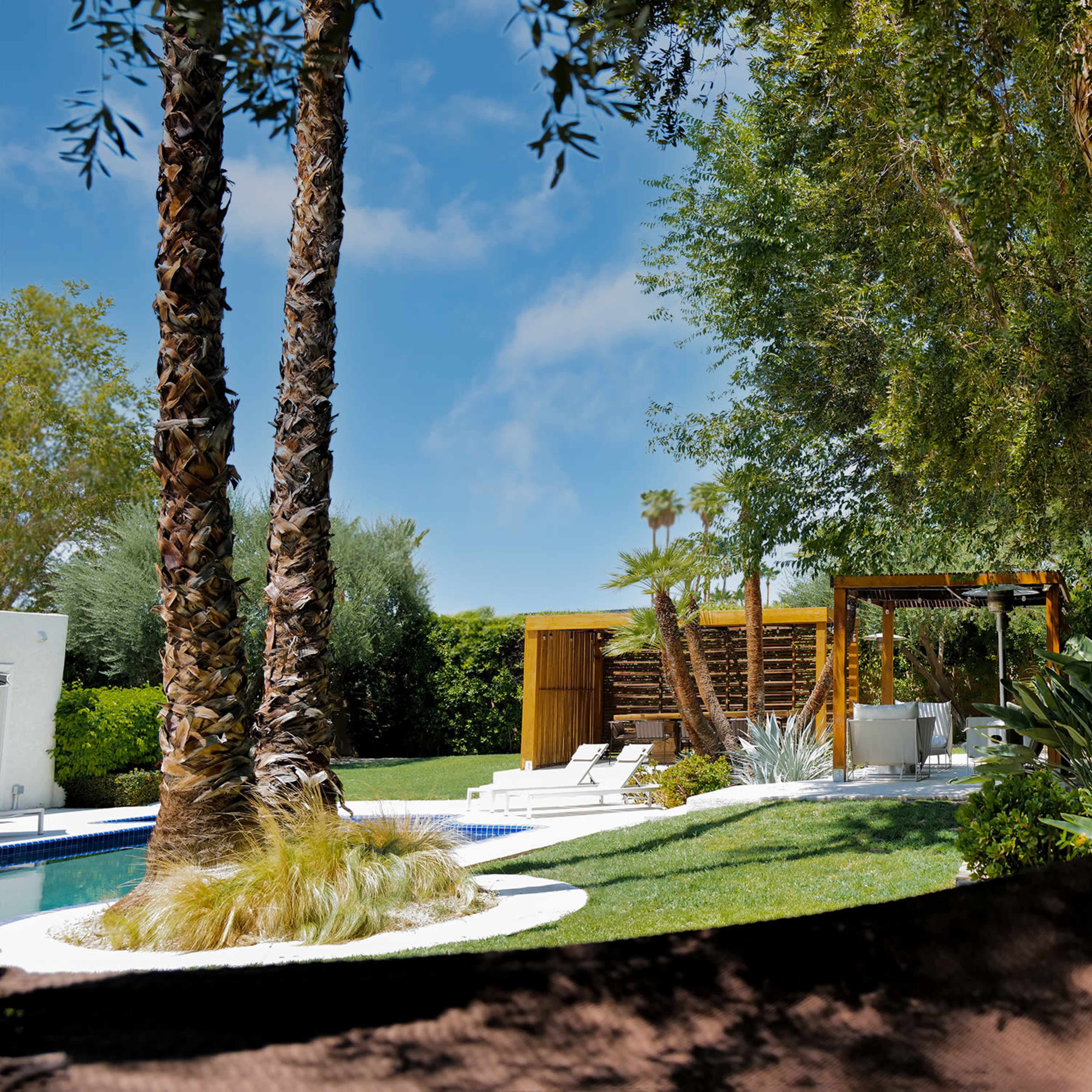 A well-maintained backyard features a pool, lounge chairs, and a shaded pergola surrounded by palm and leafy trees.
