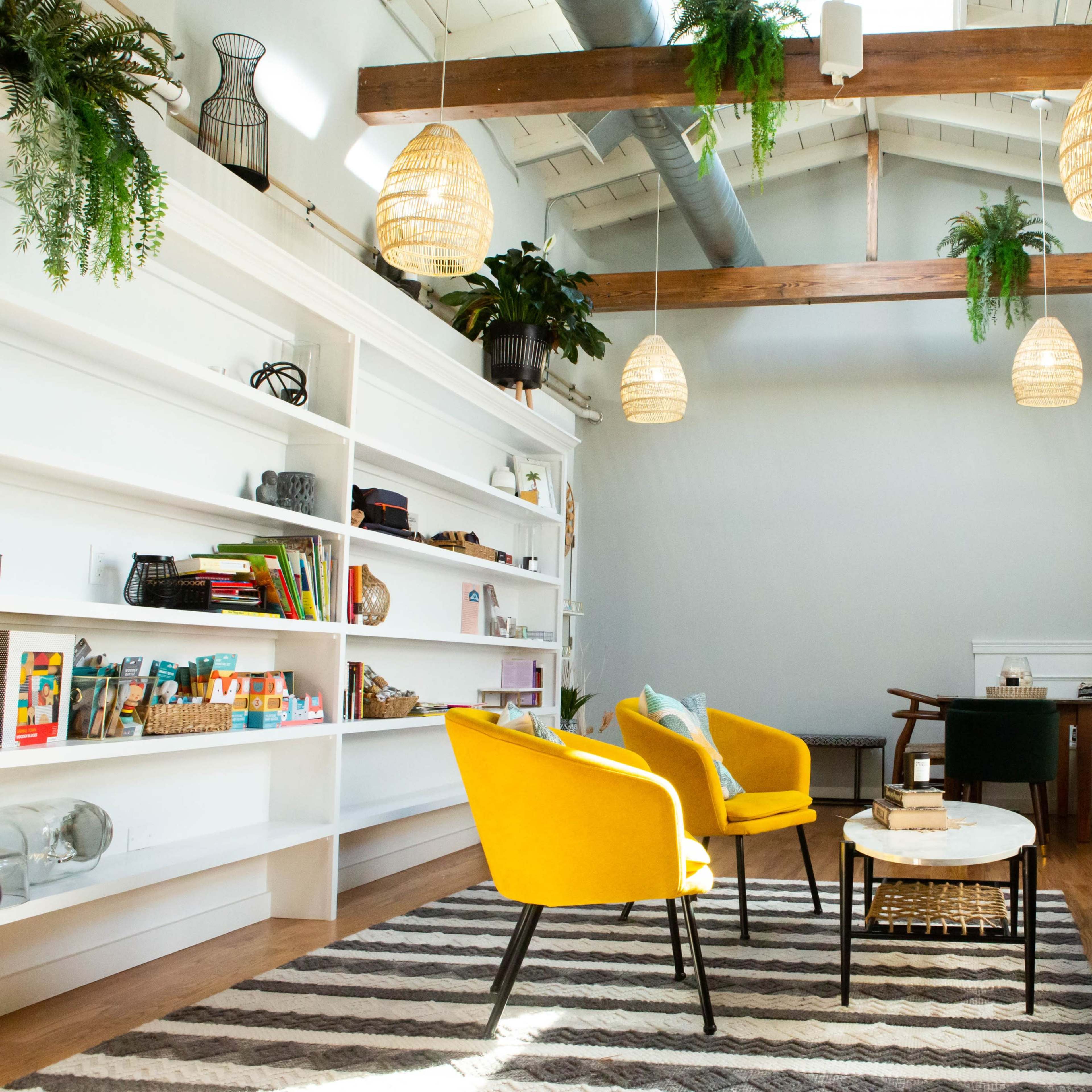 A modern living space featuring yellow armchairs, a white shelving unit filled with items, and a seating area with a coffee table on a striped rug under hanging lights.