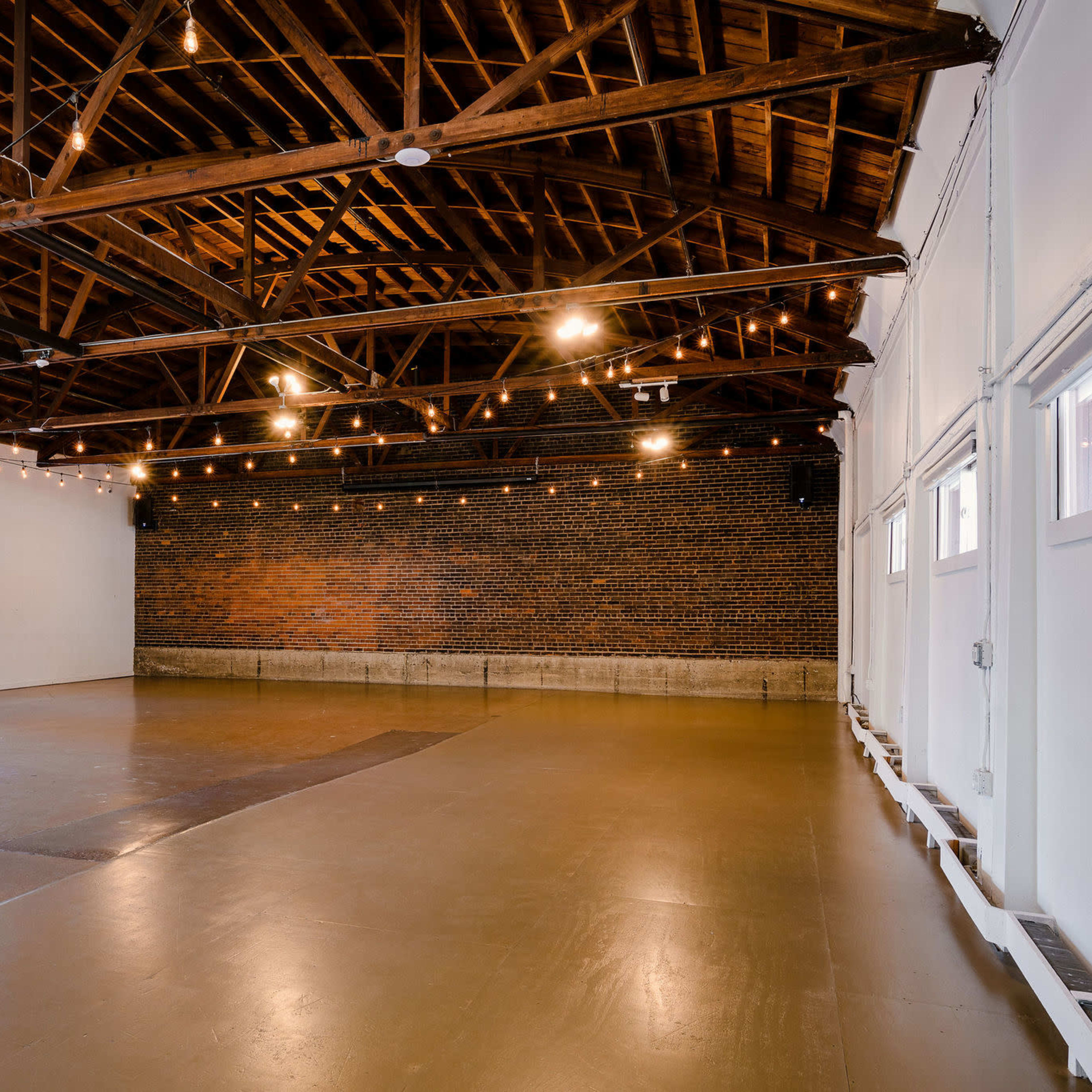 A spacious, industrial-style room with exposed wooden beams, string lights, and a brick wall, featuring a polished concrete floor and large garage doors.