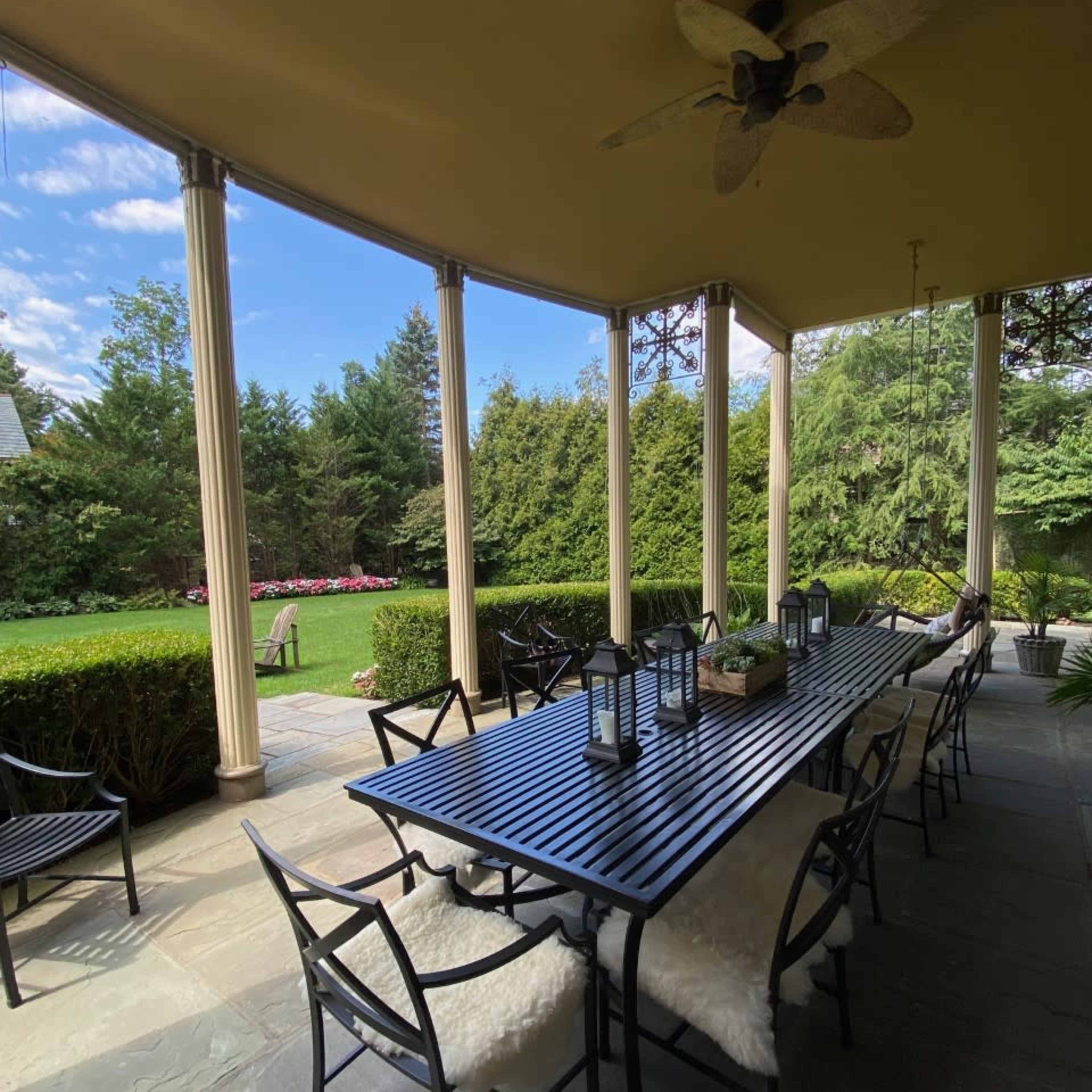 A spacious outdoor dining area is shown with a long table surrounded by chairs on a stone patio, set against a backdrop of greenery and trees.