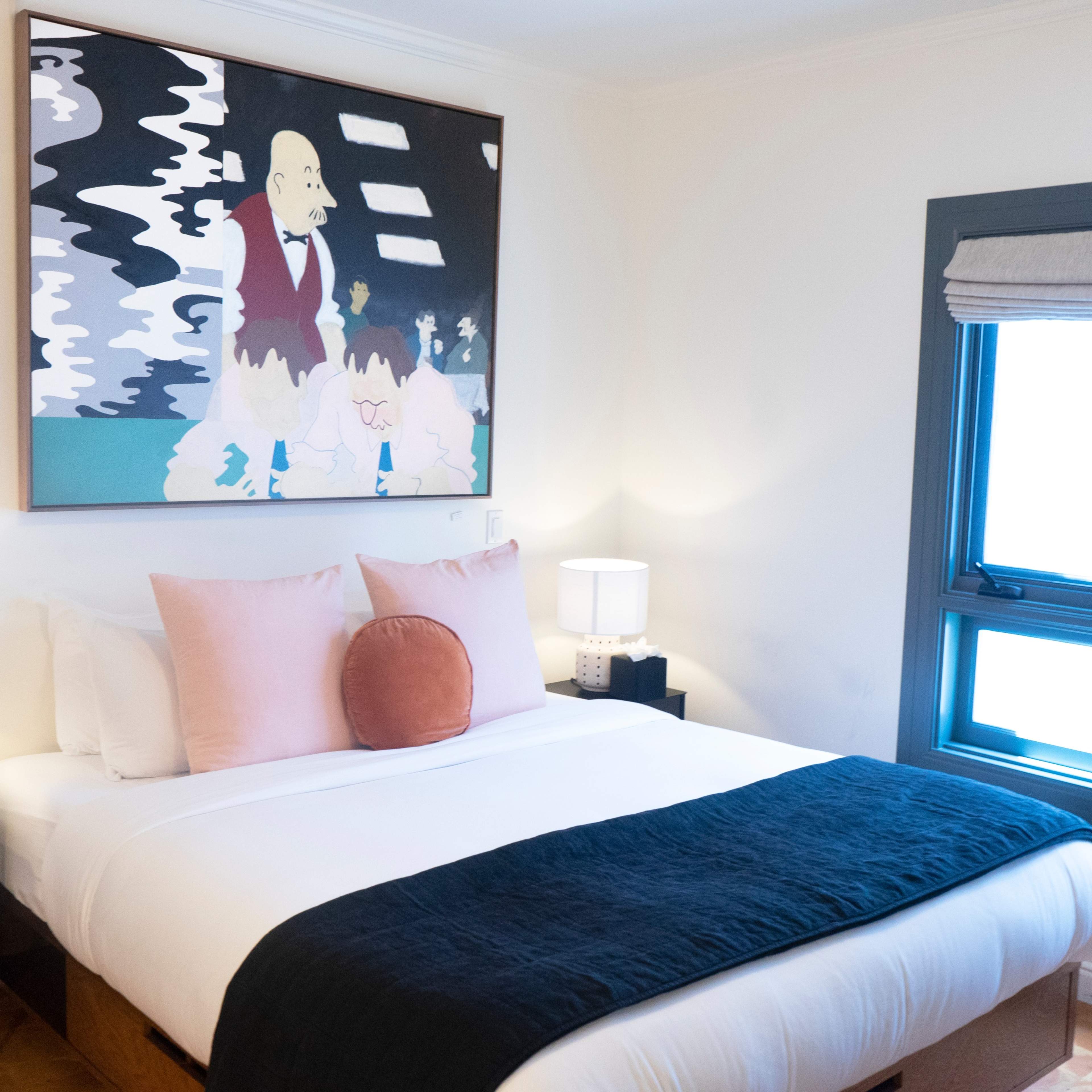 A cozy bedroom featuring a large bed with decorative pillows, two bedside lamps, and a colorful artwork above the bed, along with a window on the right side.