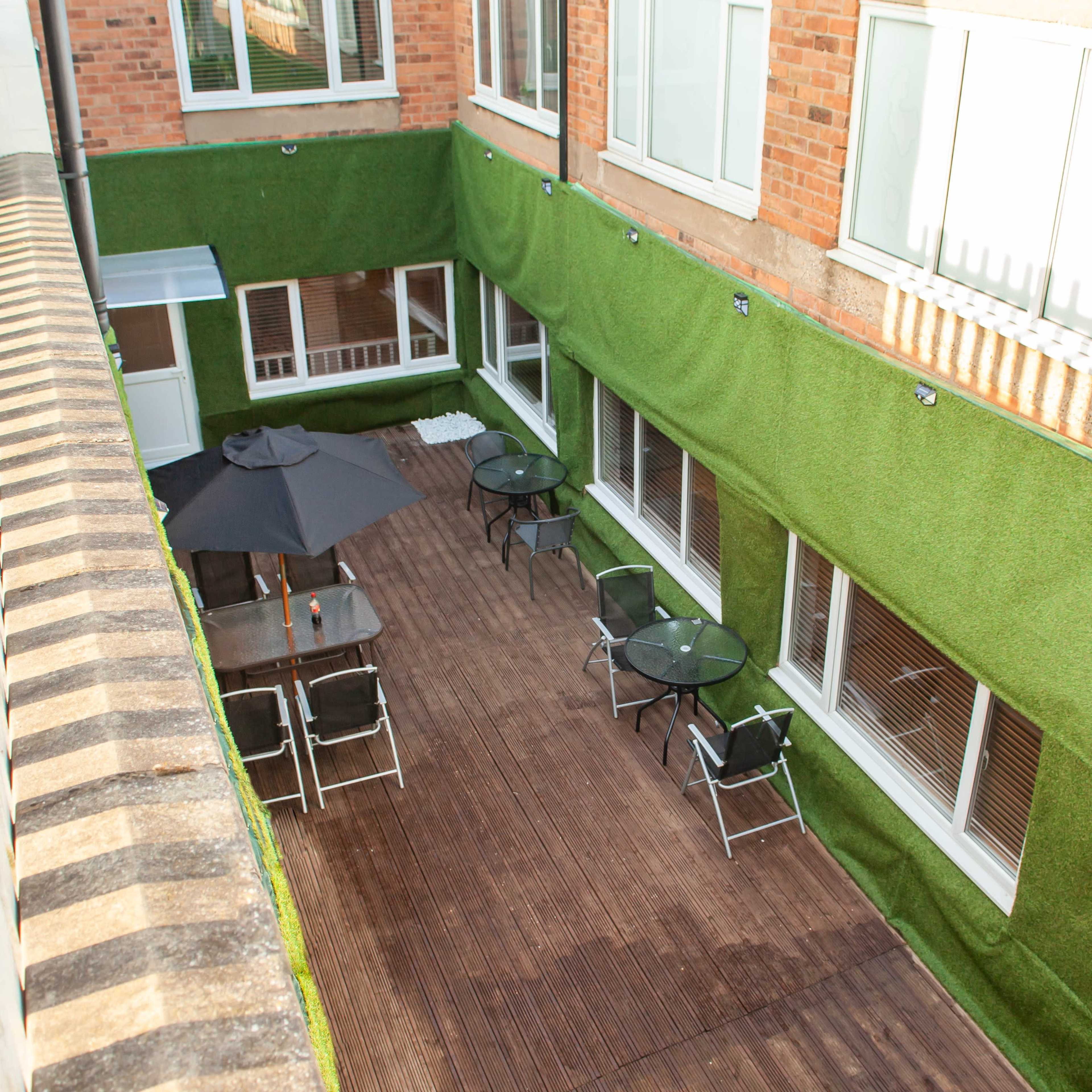 A courtyard with wooden decking, glass tables, chairs, and a large umbrella, surrounded by green artificial grass on the walls.