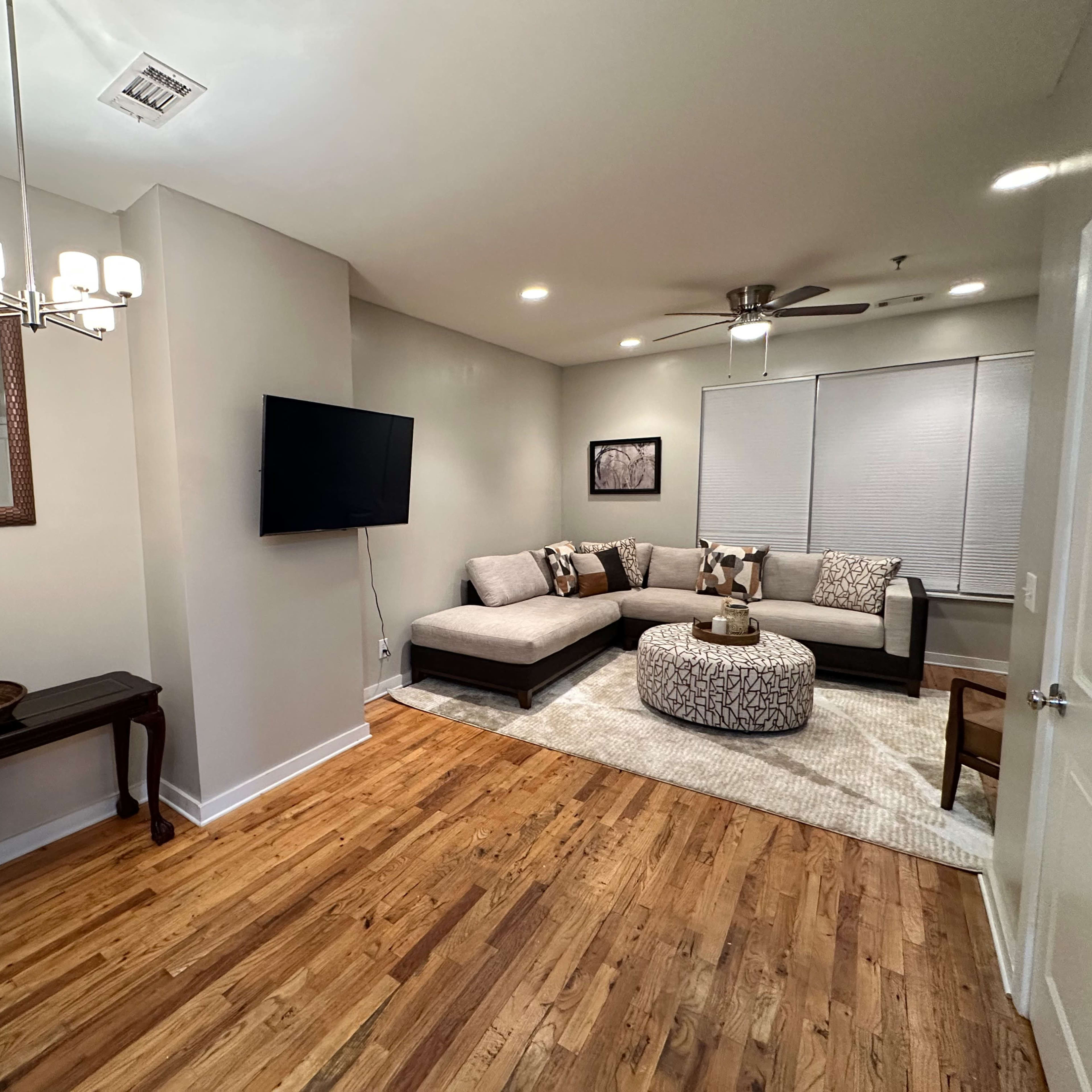 A living room featuring a sectional sofa, a coffee table with decorative items, a wall-mounted television, and wooden flooring.