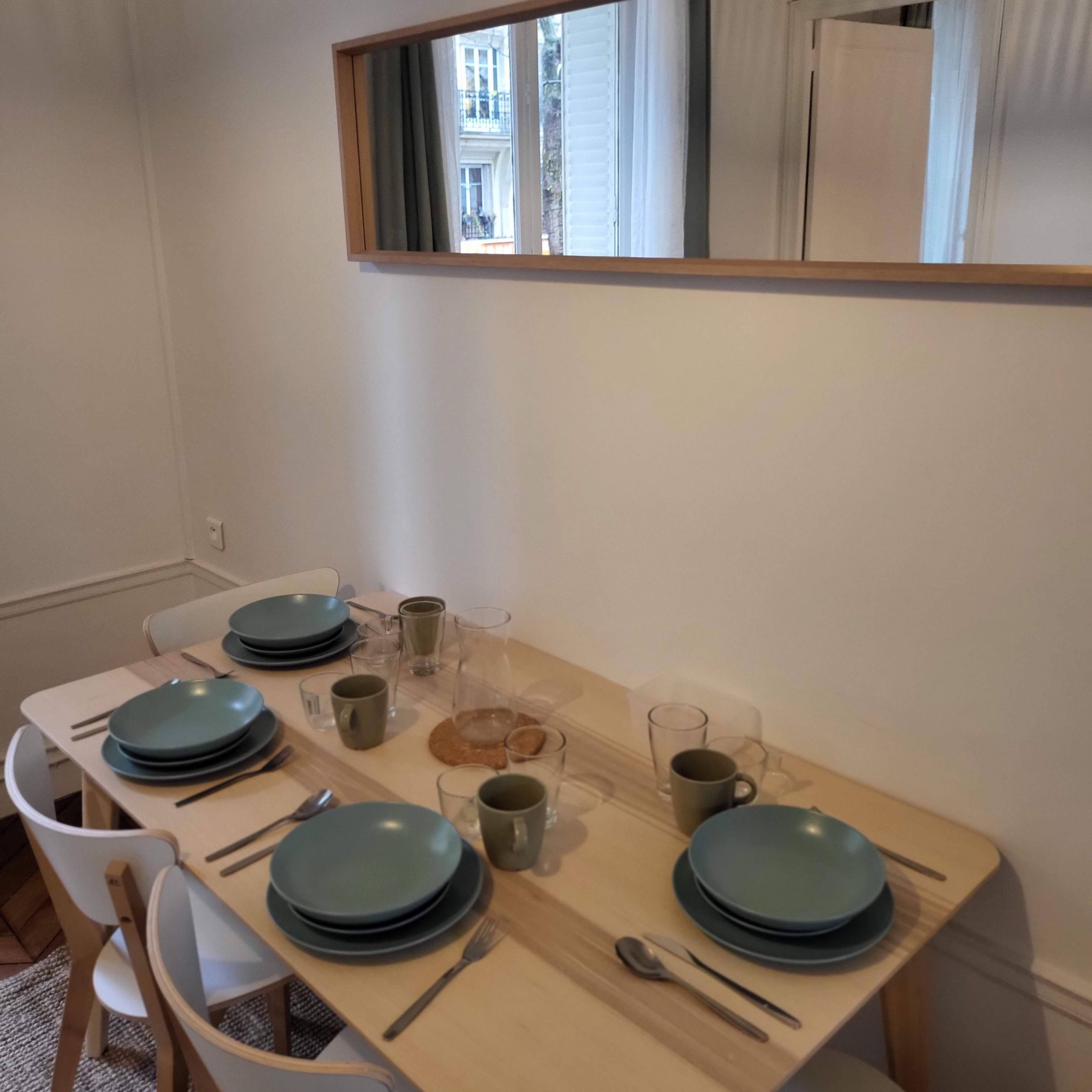 A dining table is set with plates, glasses, and utensils, reflecting light from a mirror on the wall.