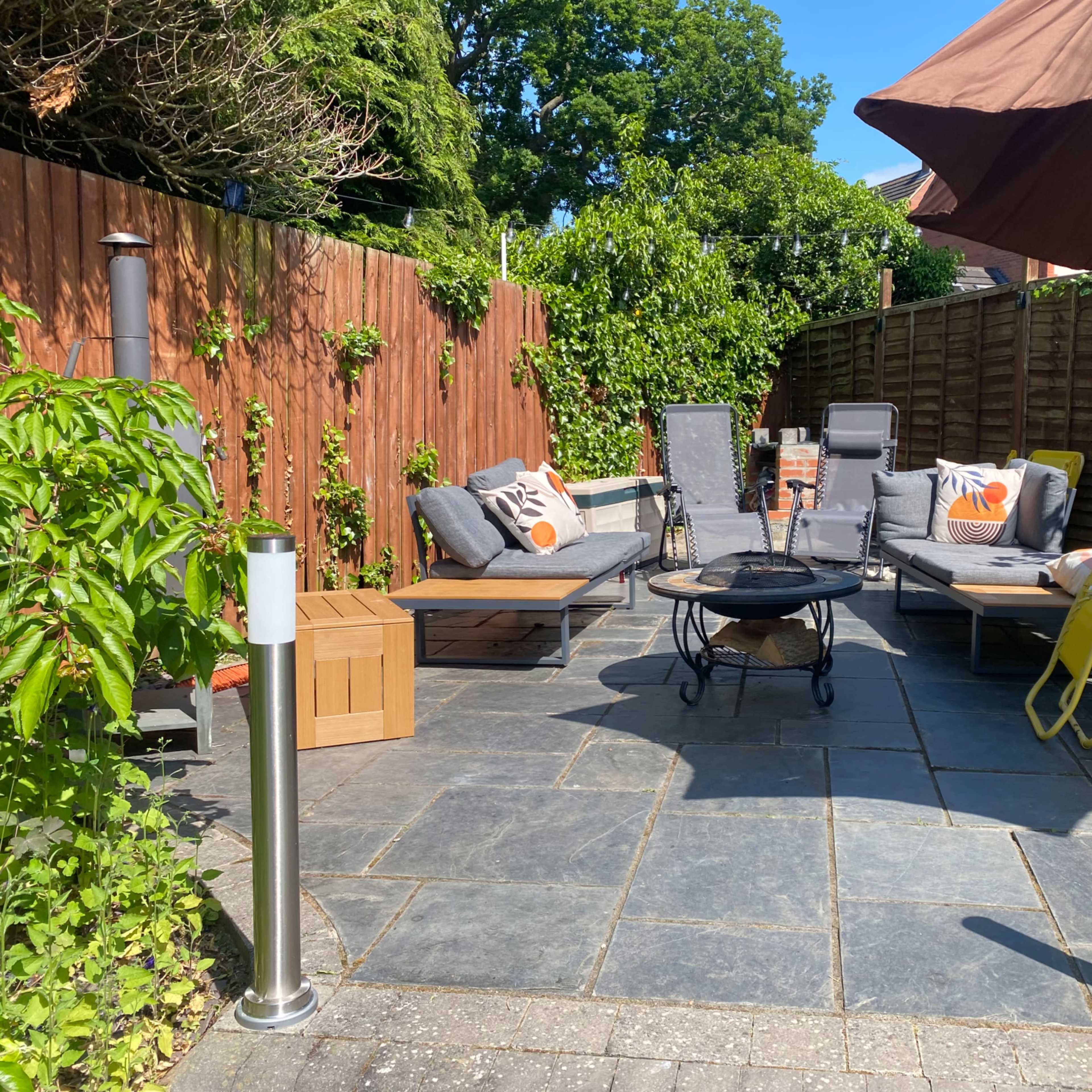 A stone-paved outdoor seating area with lounge chairs, a fire pit, and an umbrella surrounded by wooden fencing and greenery.
