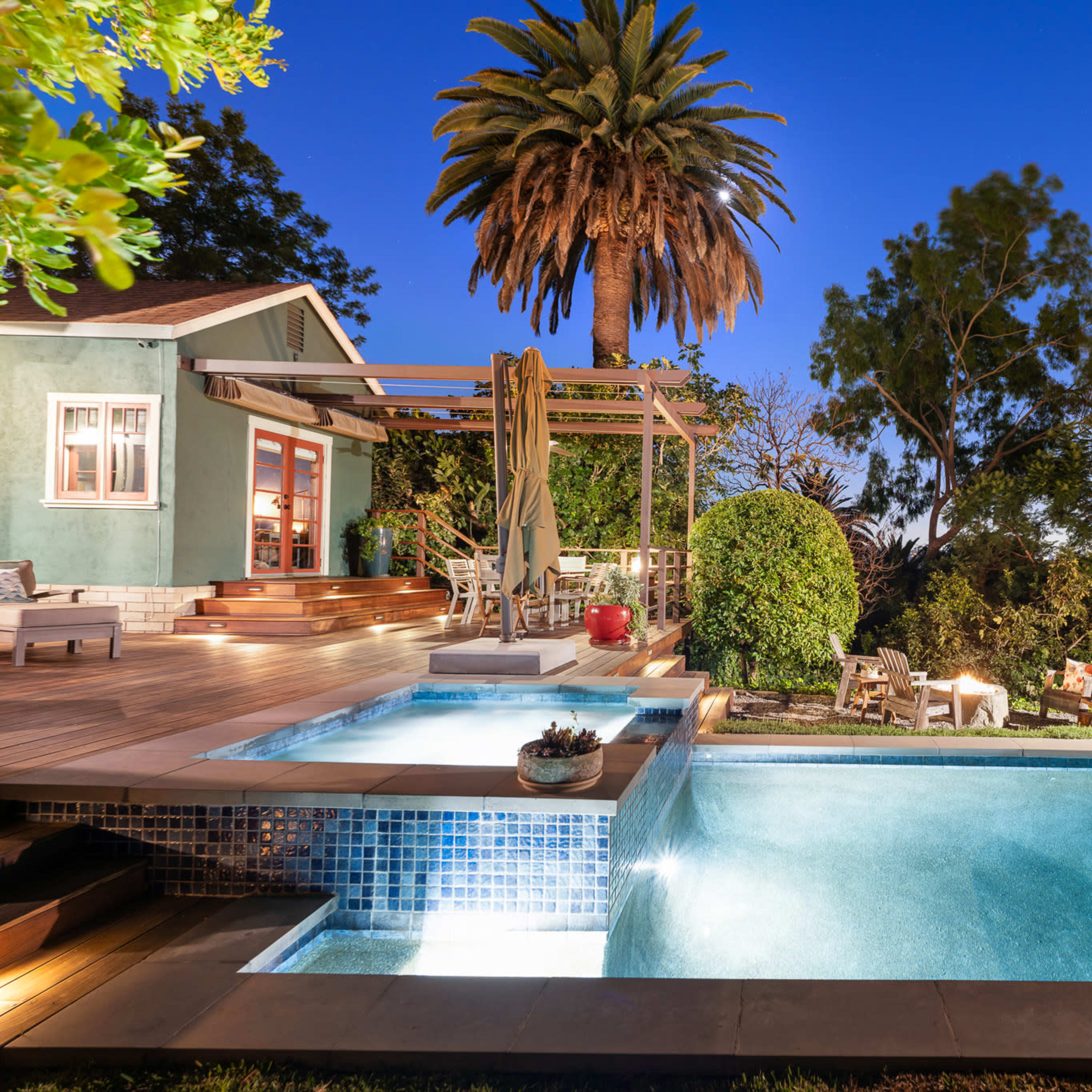 A modern home features a well-lit swimming pool surrounded by palm trees and a wooden deck at dusk.