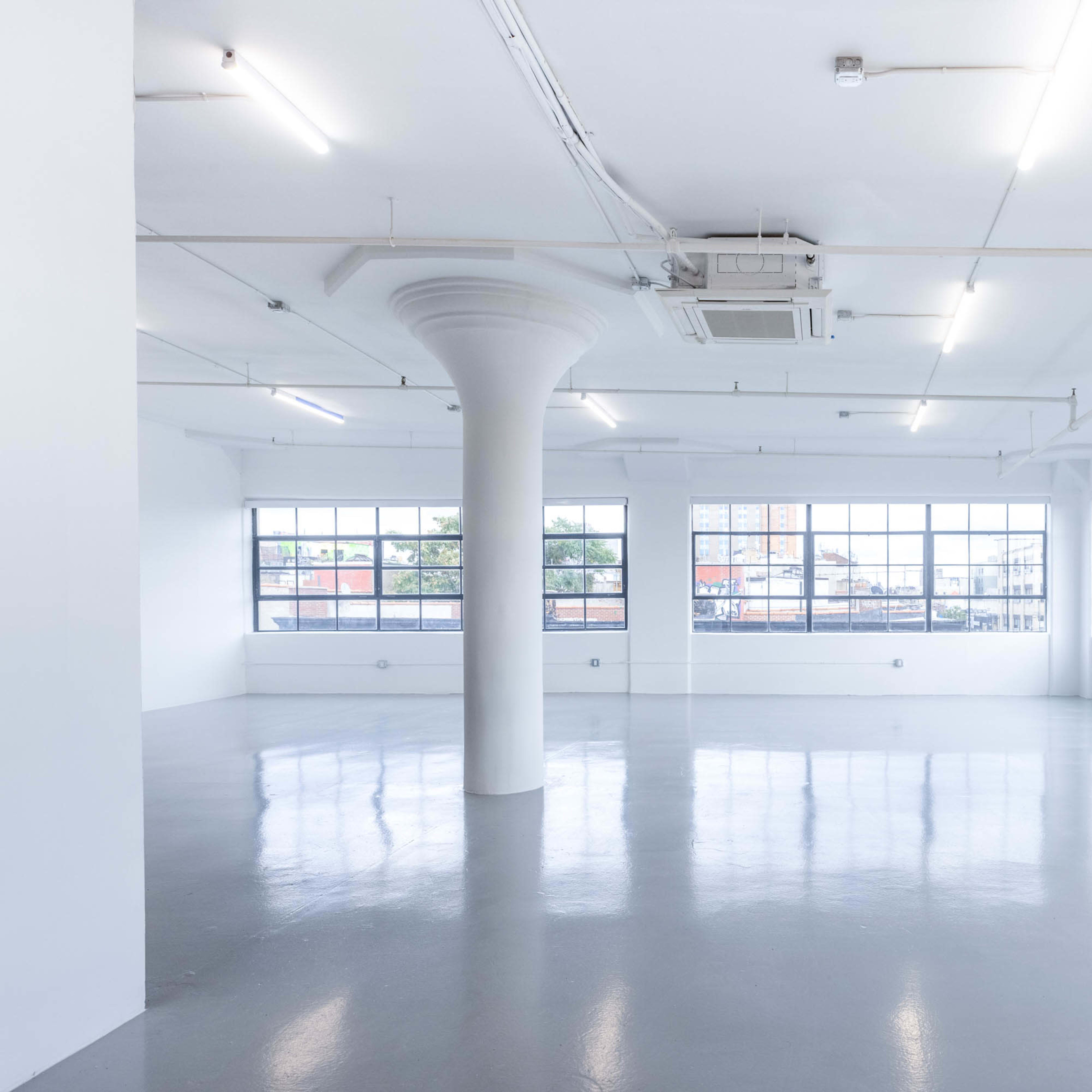 A spacious, empty room with white walls, polished gray flooring, and large windows allowing natural light to enter.