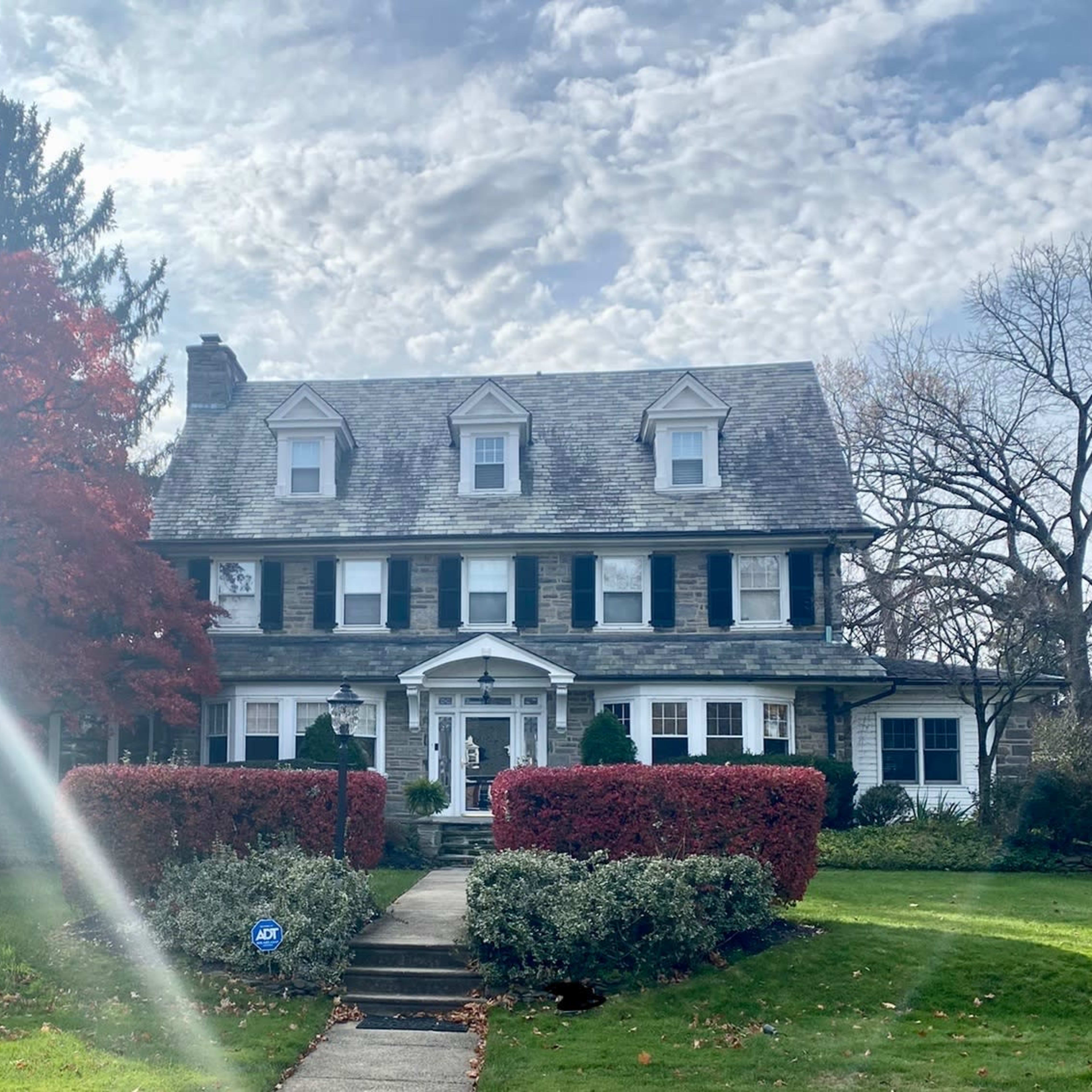 A large, gray-stone house with a gabled roof and white trim is surrounded by neatly trimmed bushes and autumn-colored trees under a cloudy sky.
