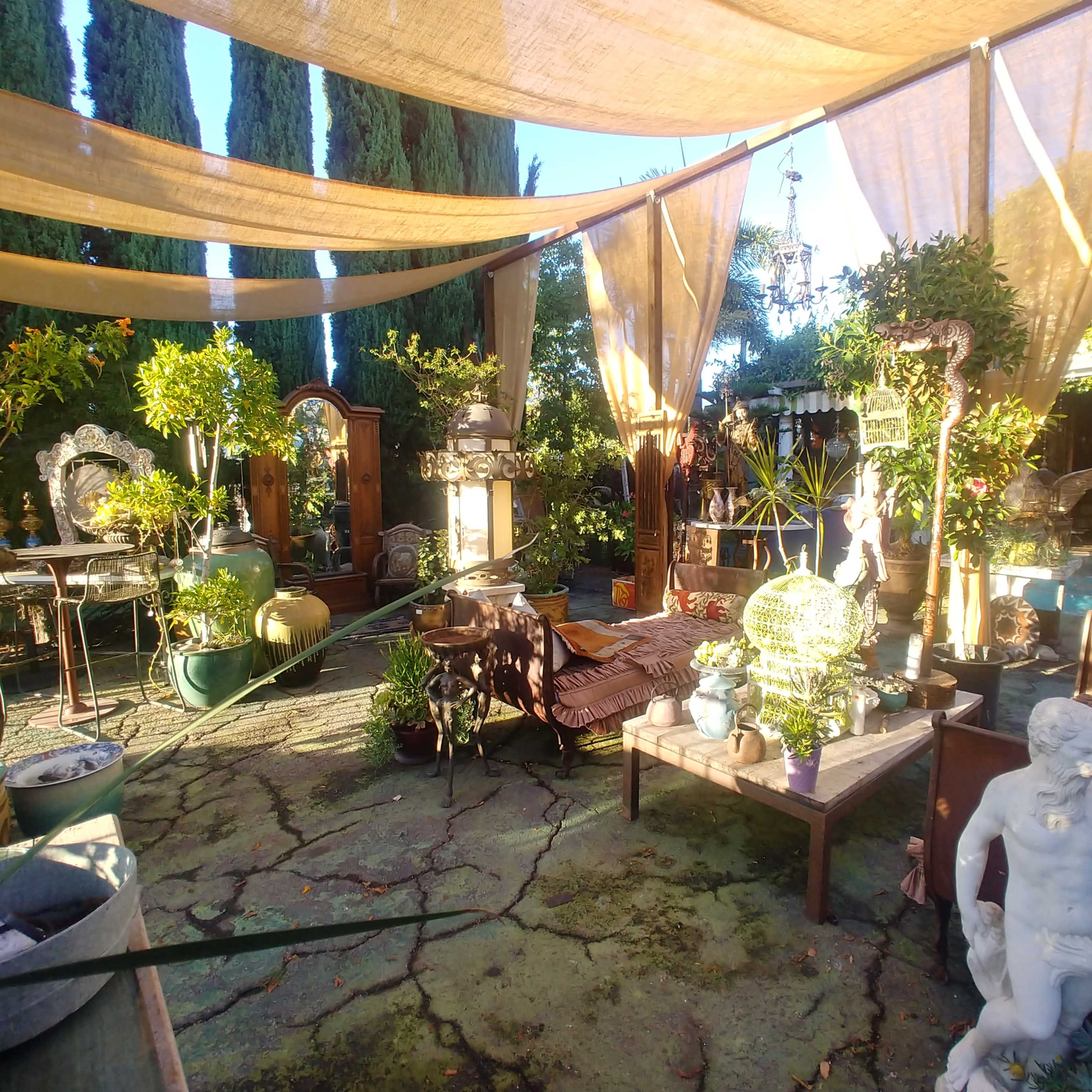 A tranquil outdoor space features various plants, sculptures, and furniture under fabric canopies.