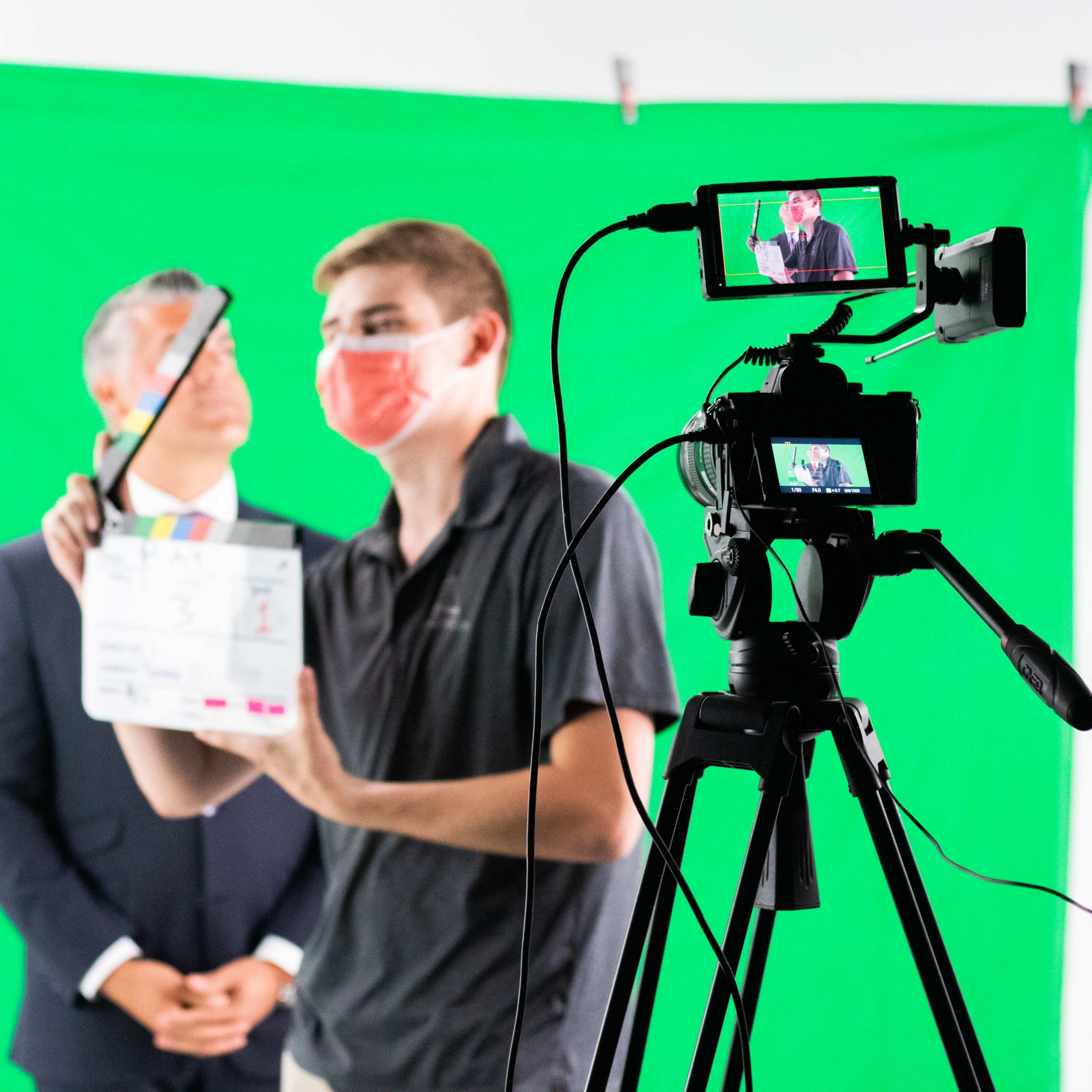 A person wearing a mask operates a camera while holding a clapperboard in front of a man in a suit against a green screen backdrop.
