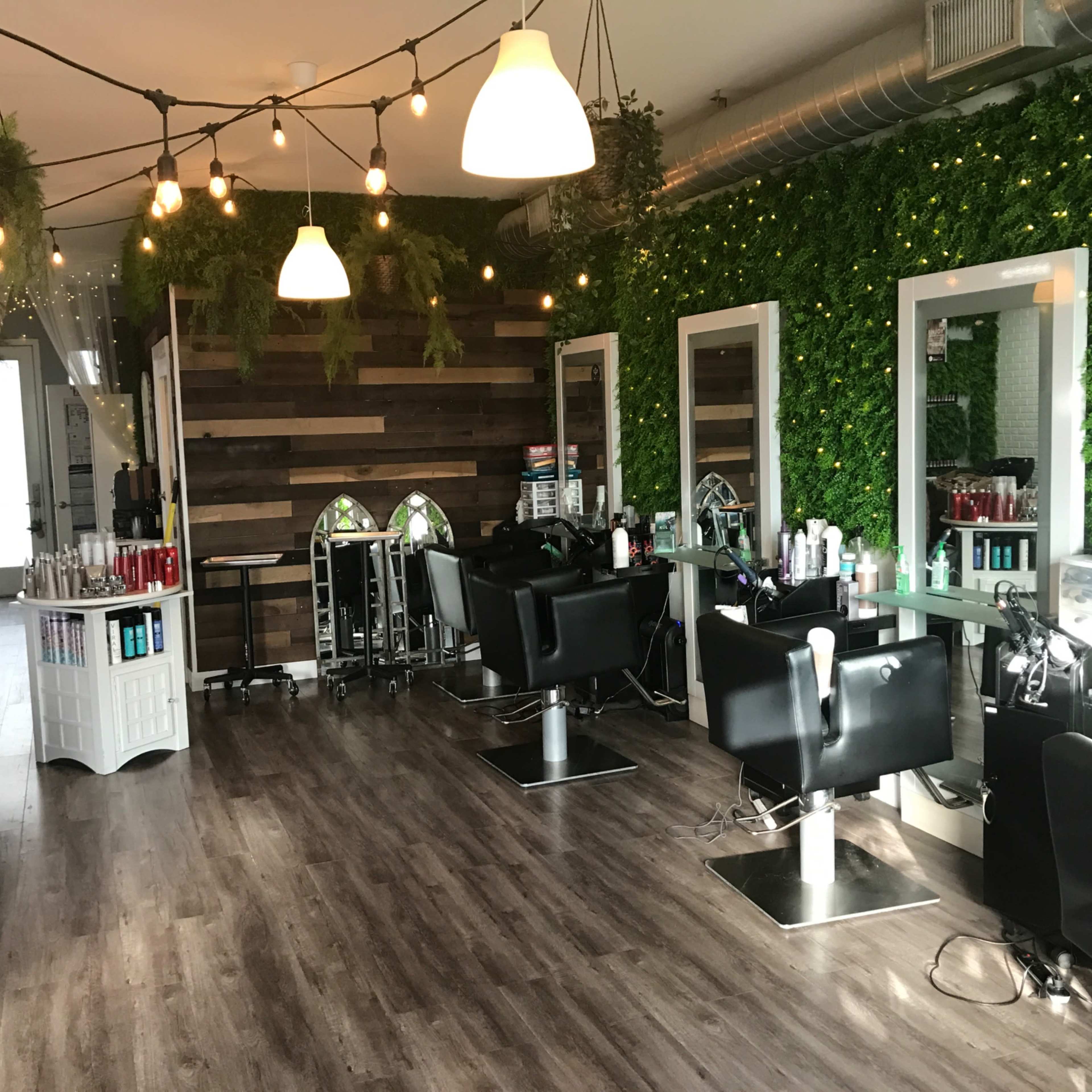 A salon interior featuring black styling chairs, mirrored stations, and greenery on the walls, with overhead pendant lights illuminating the space.