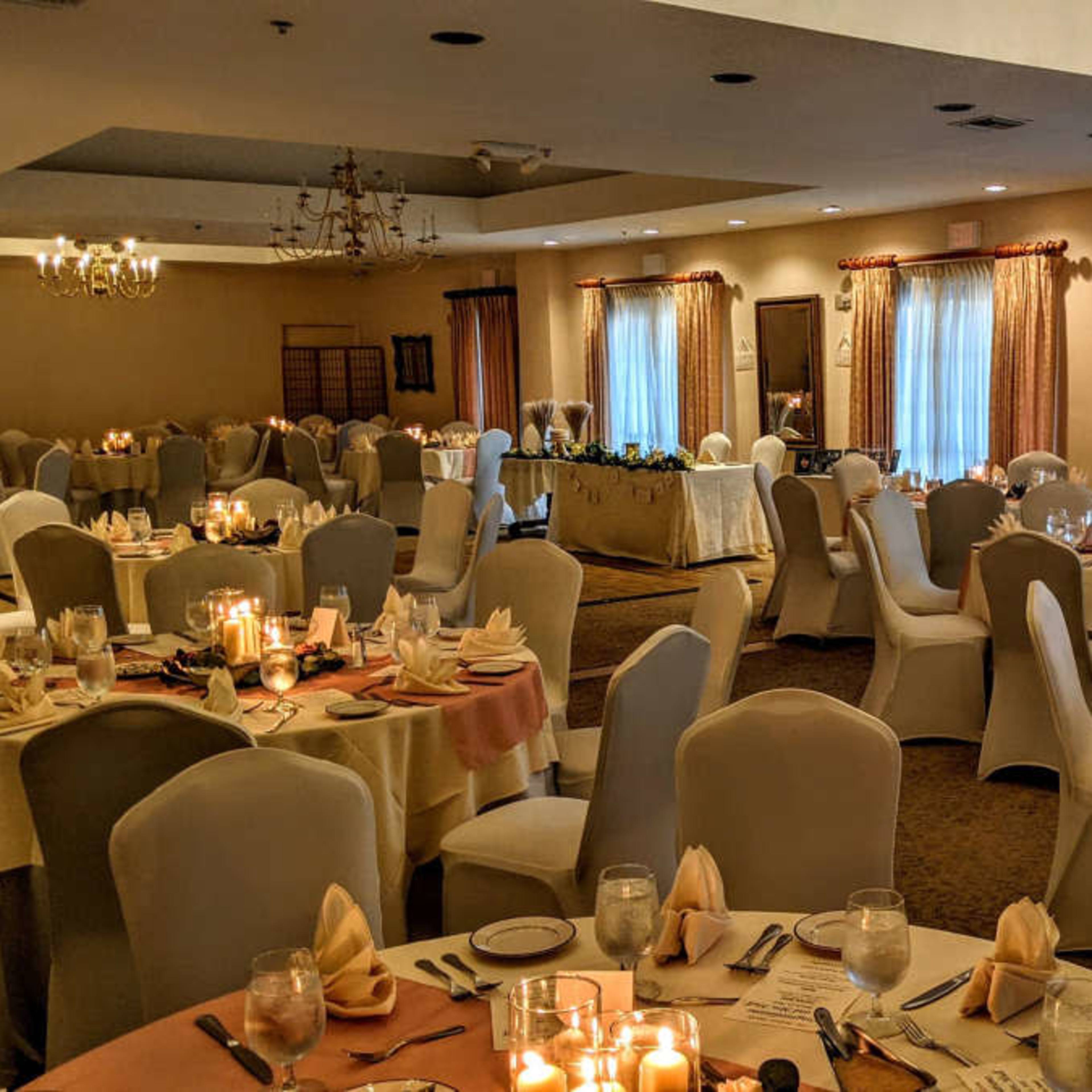 A banquet hall set for an event, featuring round tables with beige linen, neatly arranged place settings, and soft lighting from candles.