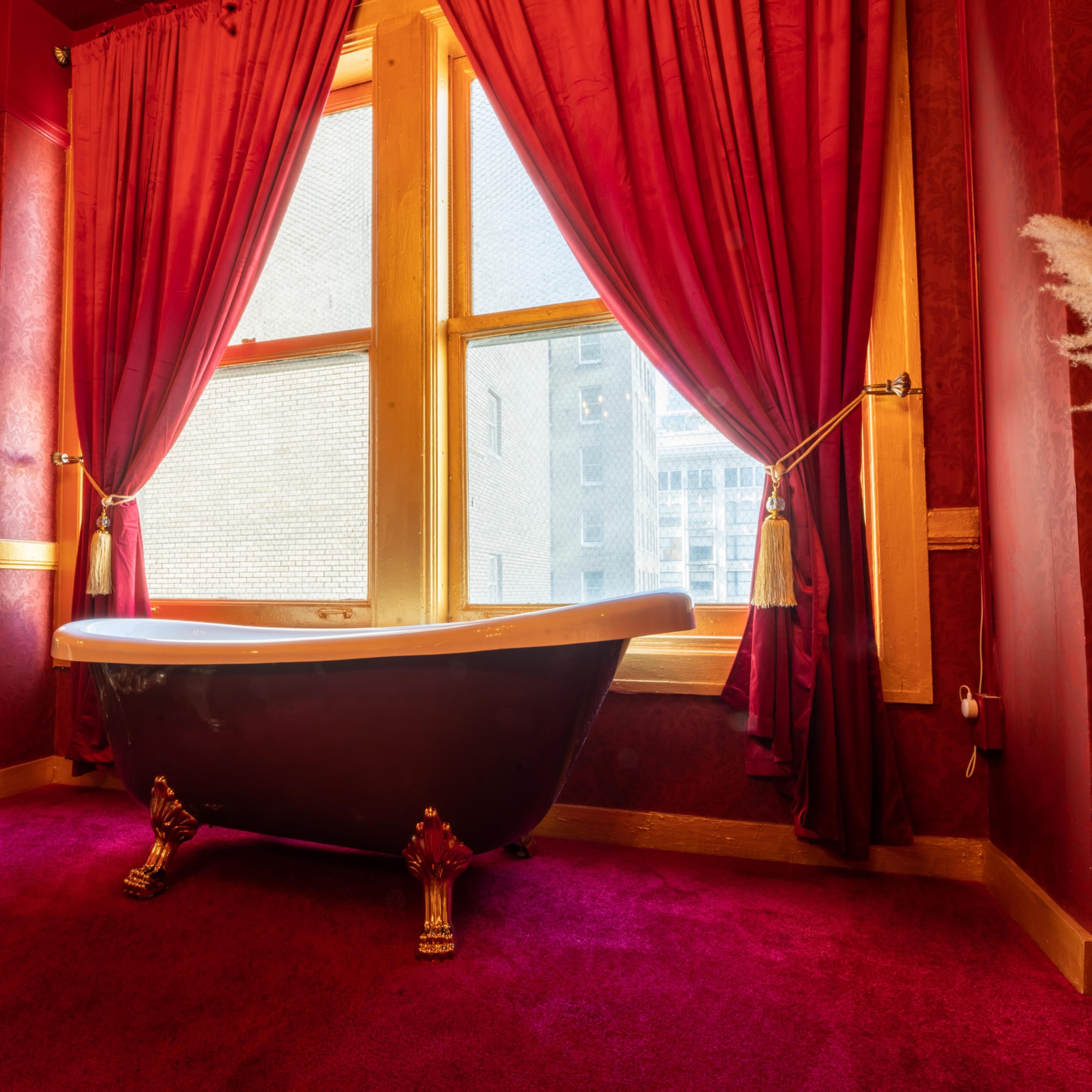 A black clawfoot bathtub is positioned in a richly decorated room with red wallpaper and curtains, featuring a plush purple carpet and a gold-accented chair.