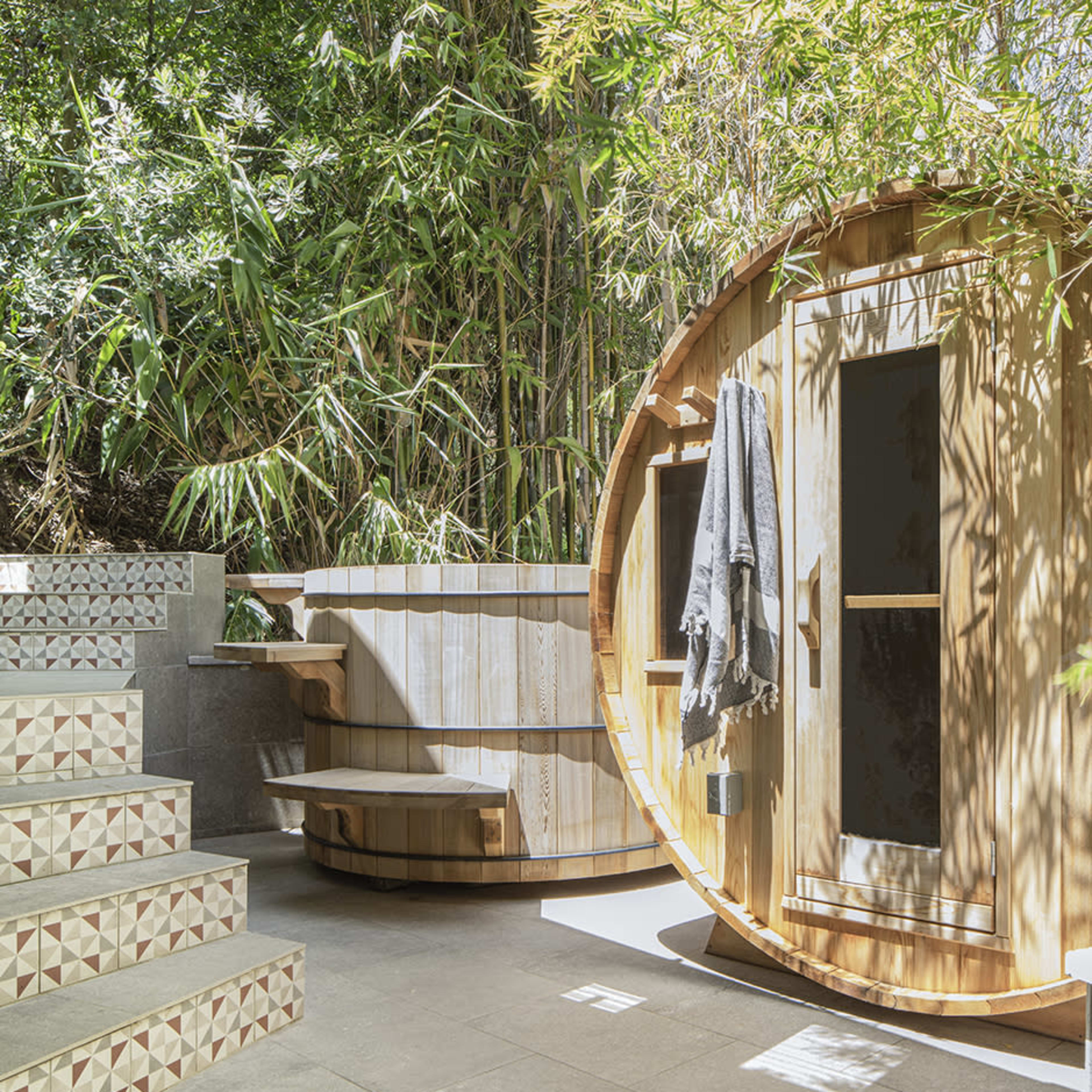 The scene features a circular wooden sauna adjacent to a round hot tub, surrounded by bamboo and stone steps.