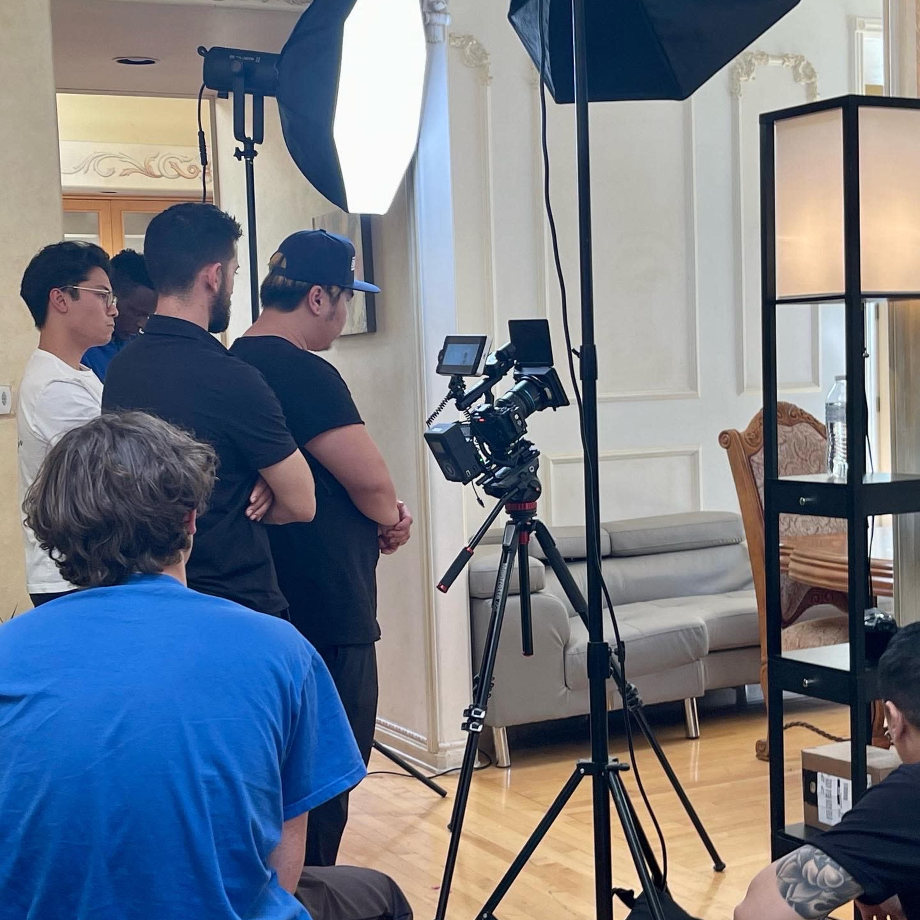 A group of people observes a camera setup with lighting equipment in a well-lit room.
