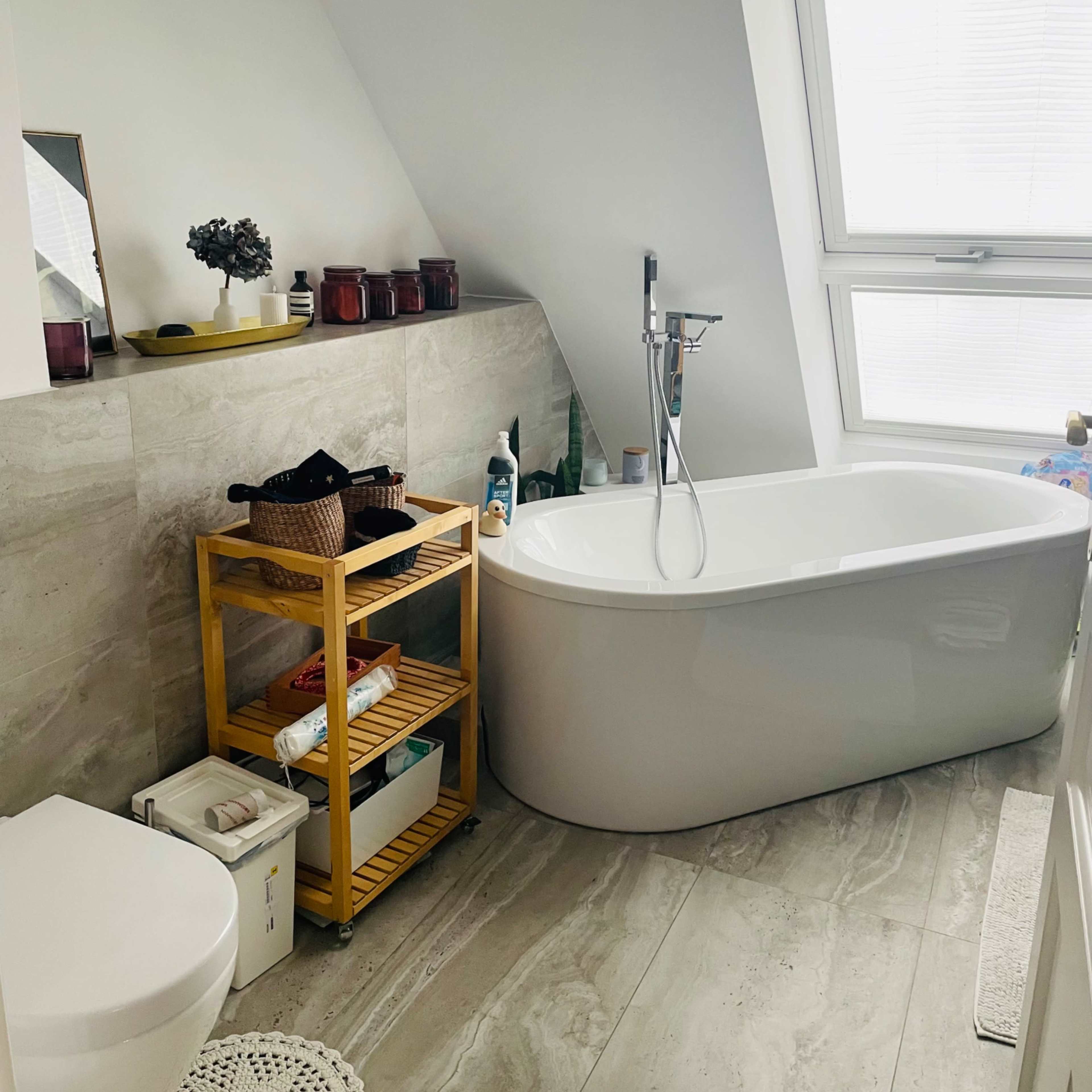 The bathroom features a freestanding bathtub, a modern sink area, a wooden storage cart, and a large window allowing natural light.