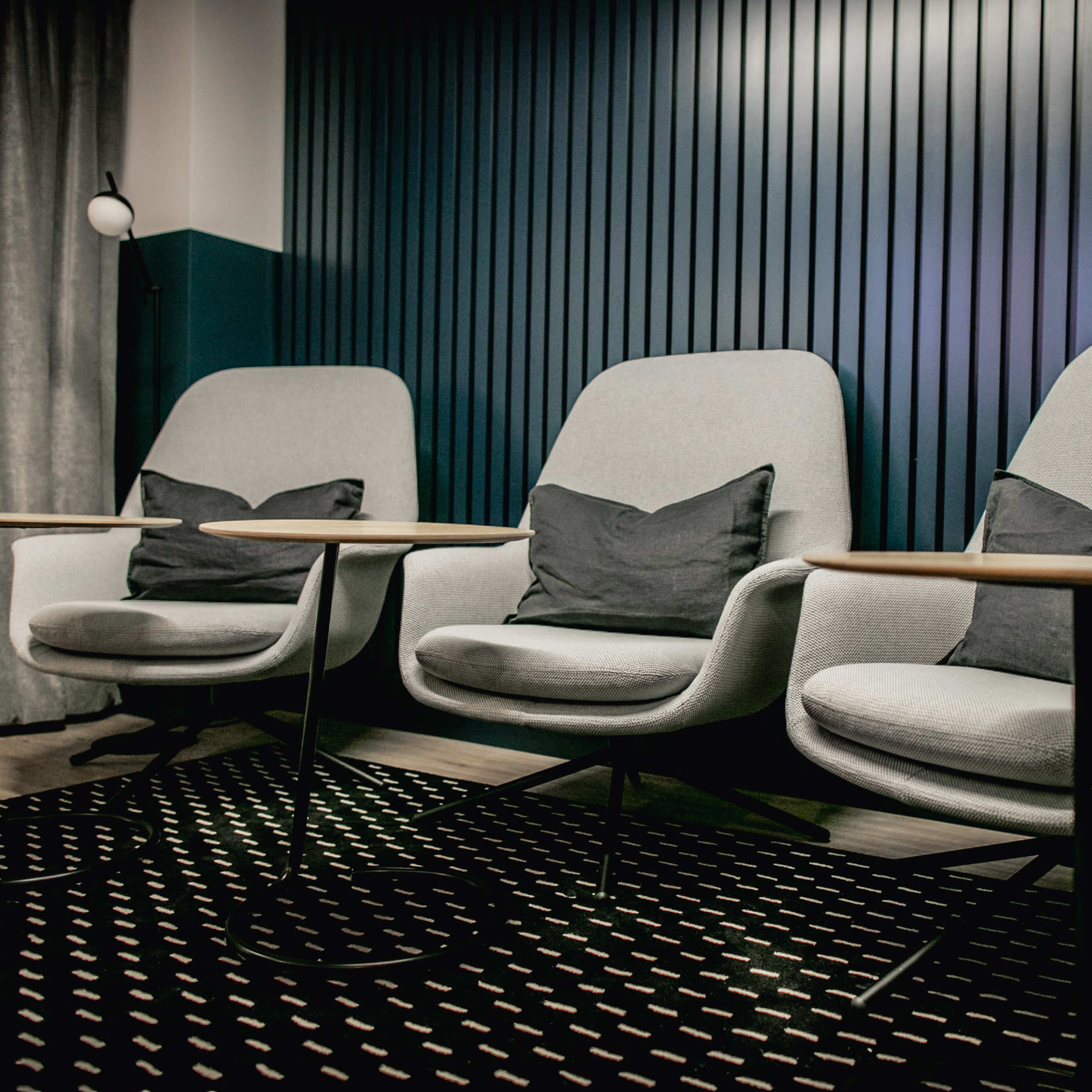 Three modern gray chairs with cushions, paired with small round wooden tables, arranged in a stylish indoor space with a textured dark wall.