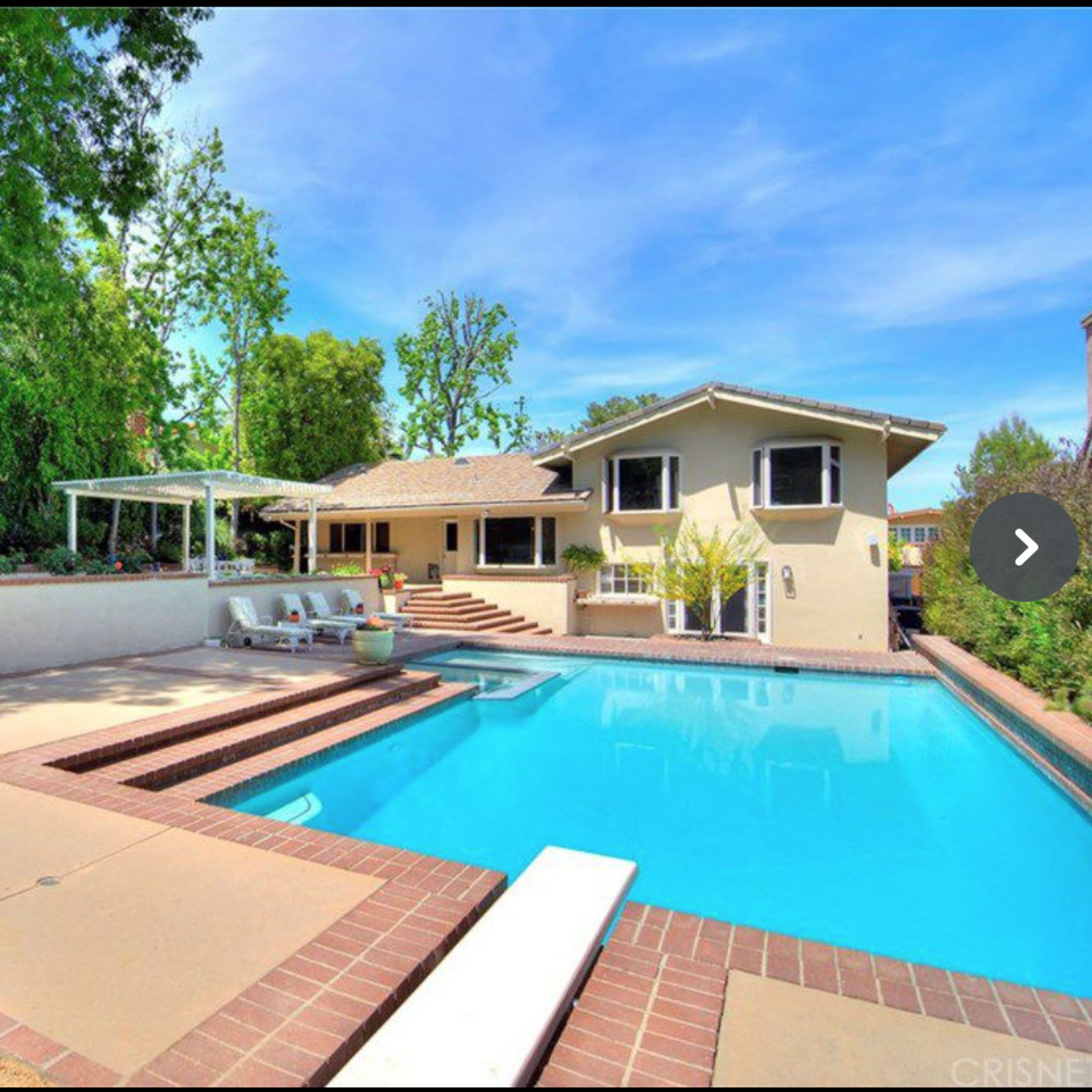 A spacious backyard featuring a swimming pool, lounge area, and a house with a patio surrounded by trees and greenery.