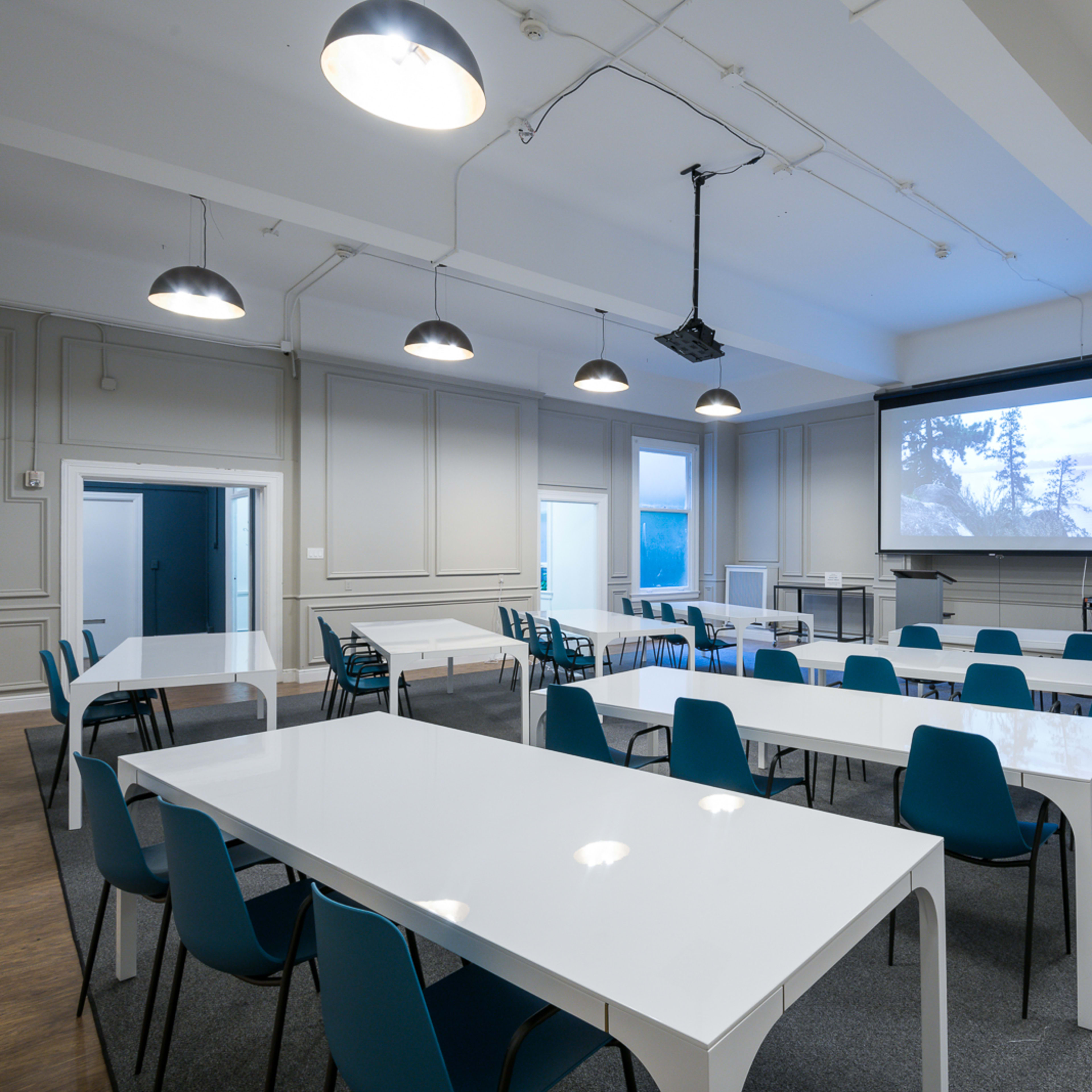 A spacious conference room features white tables arranged in a U-shape, blue chairs, and a projector screen at the front.